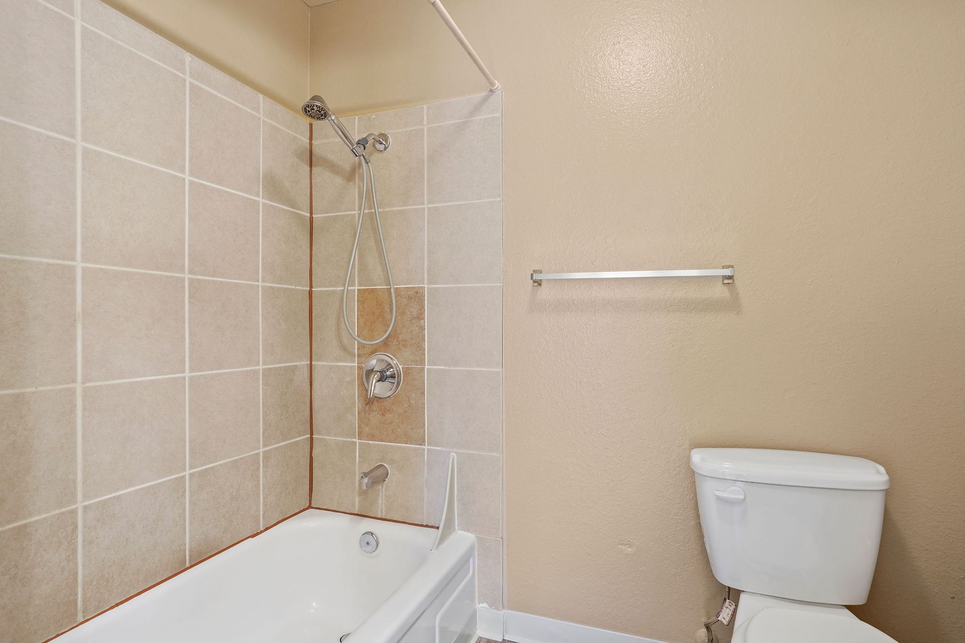 Small beige bathroom with bathtub, showerhead, towel bar, and white toilet.