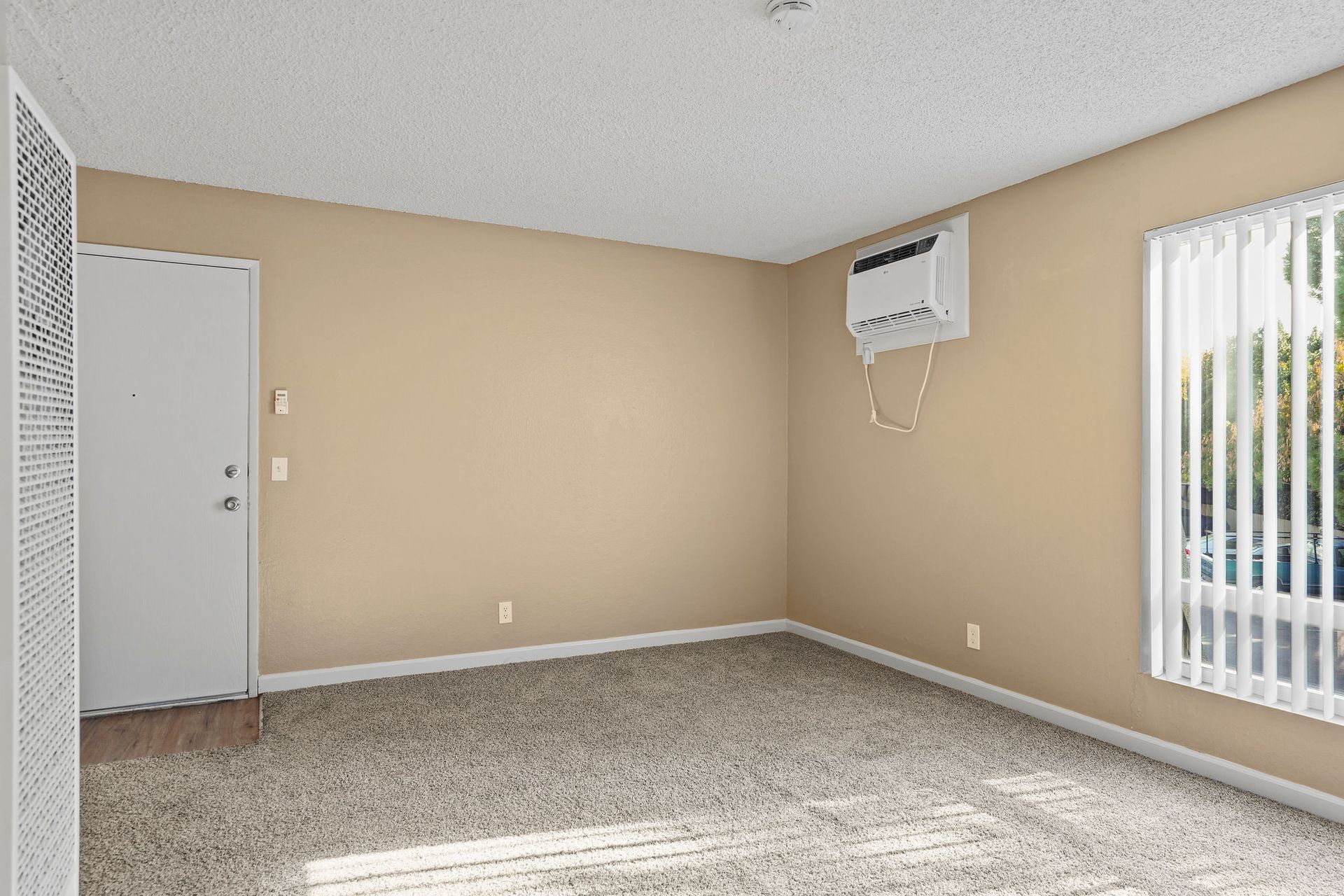 Empty beige room with carpet, white door, wall-mounted AC unit, and window blinds.