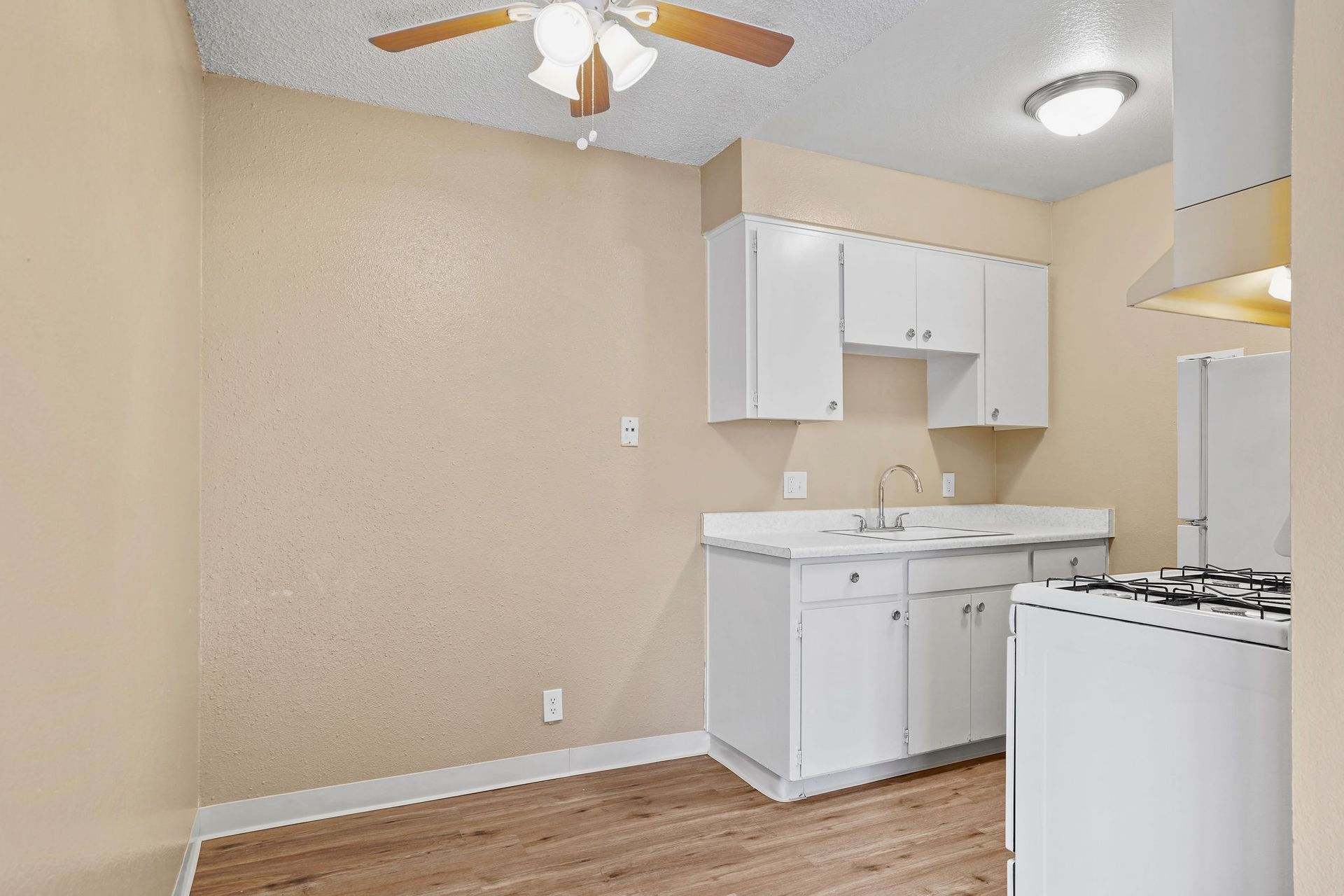Small beige kitchen with white cabinets, sink, stove, ceiling fan, and wood-look flooring.