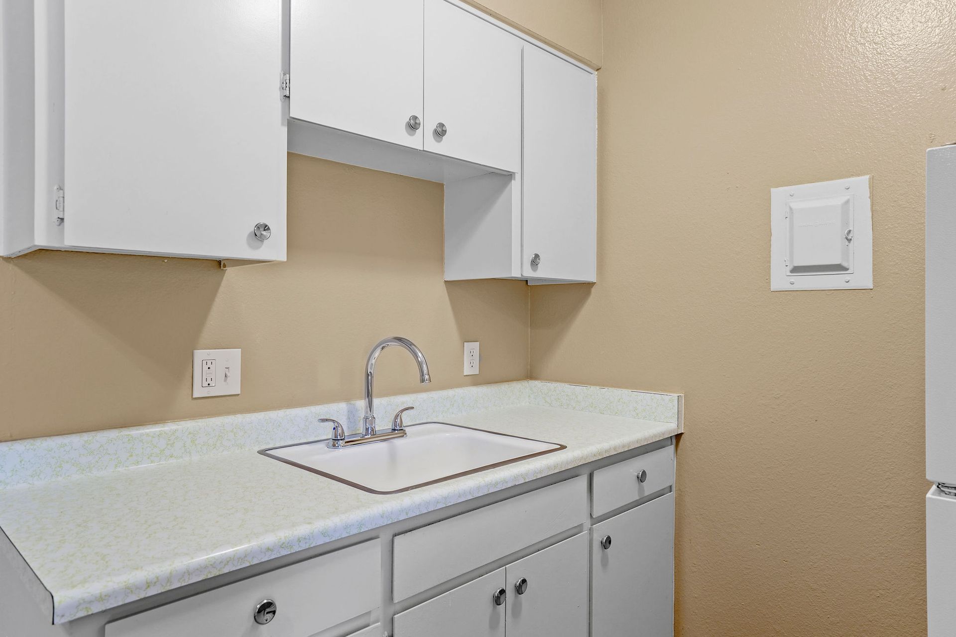 Small beige kitchen with white cabinets, sink, countertop, and wall-mounted paper towel holder.