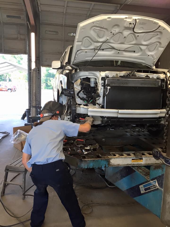 A person wearing a welding helmet works on the front engine compartment of a white truck in a repair shop.