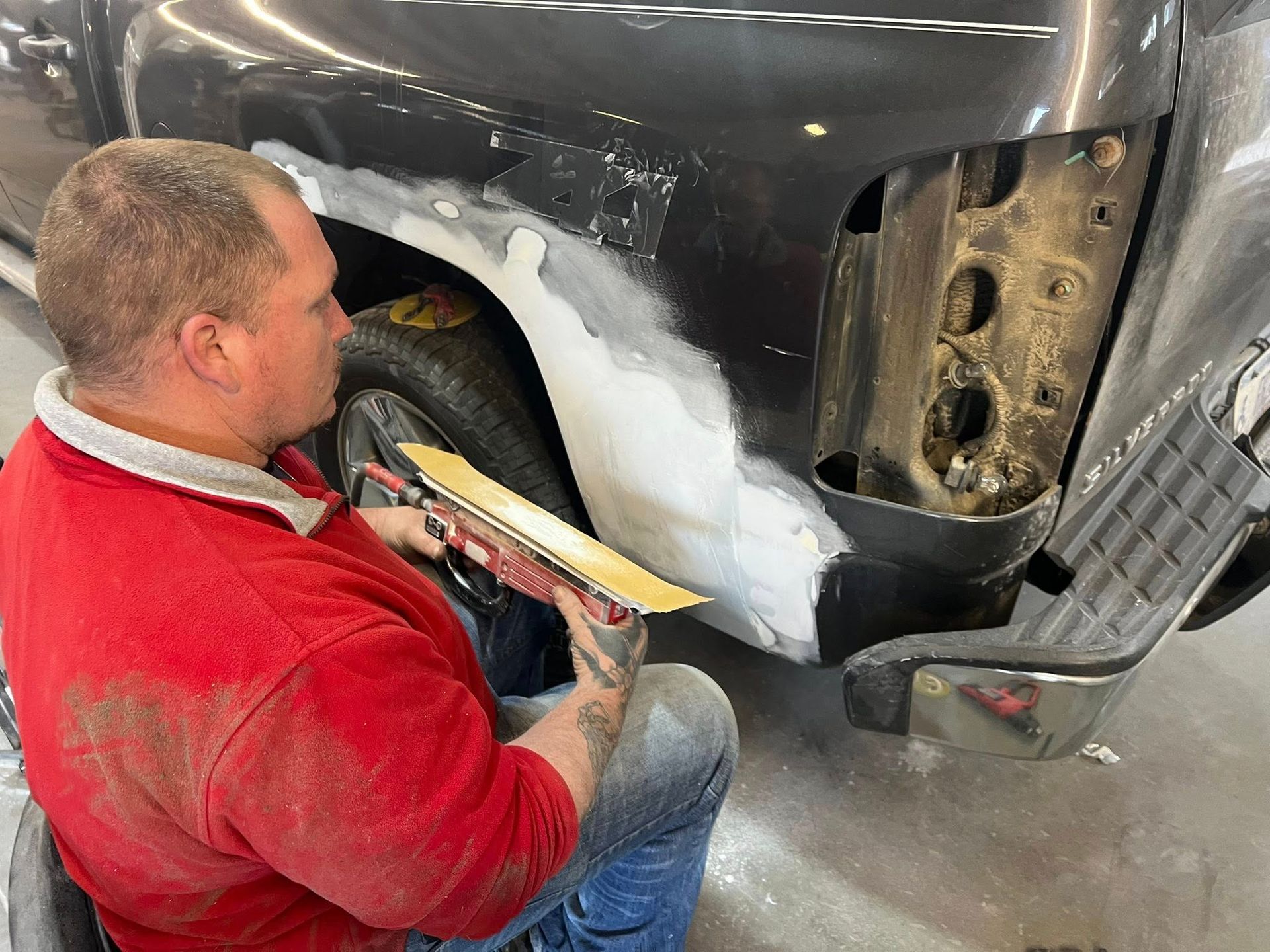 A person in a red shirt sands a white body filler patch on the rear quarter panel of a grey truck in a workshop.