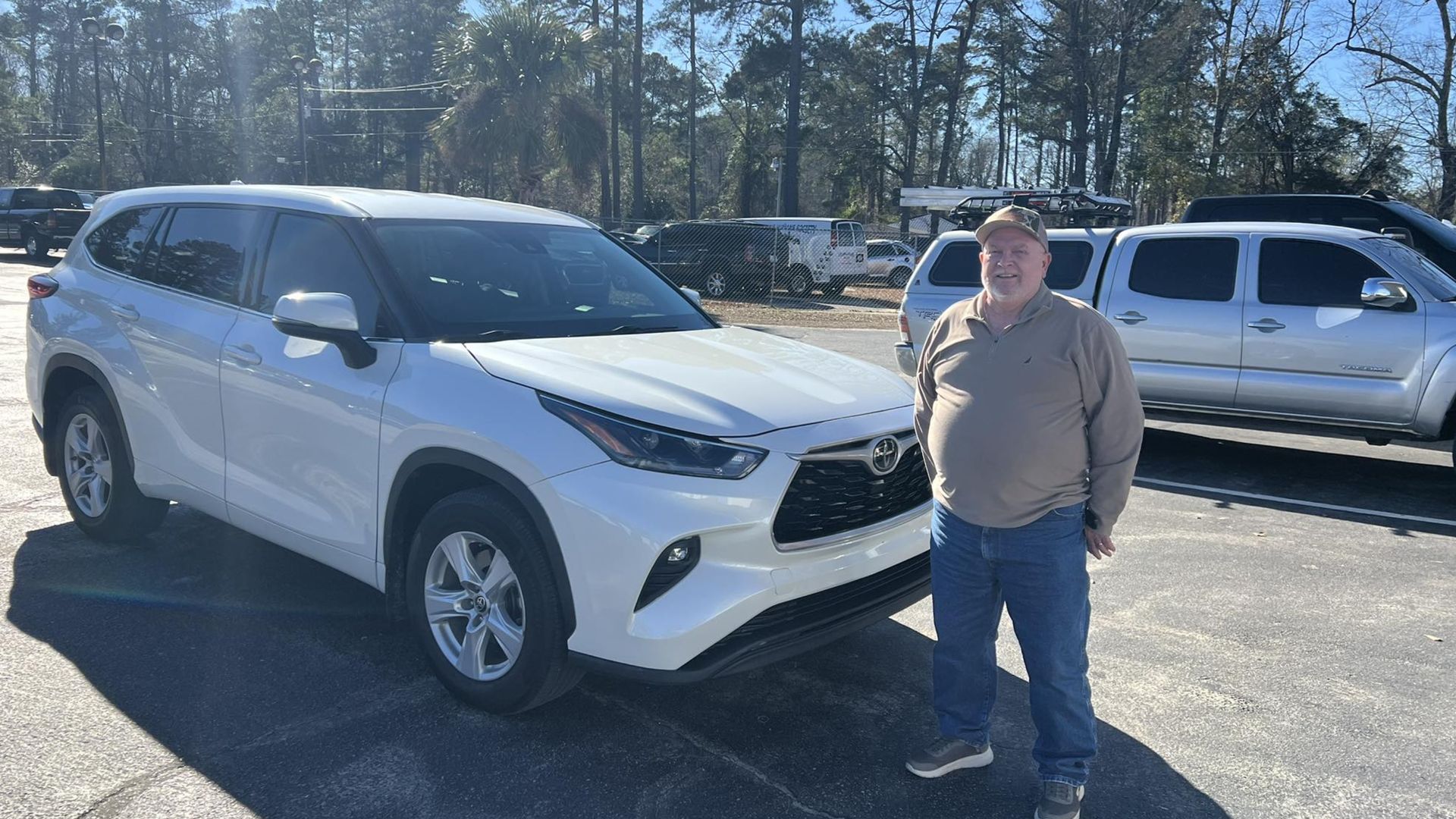 A person stands in a sunny parking lot next to a new white Toyota Highlander SUV.