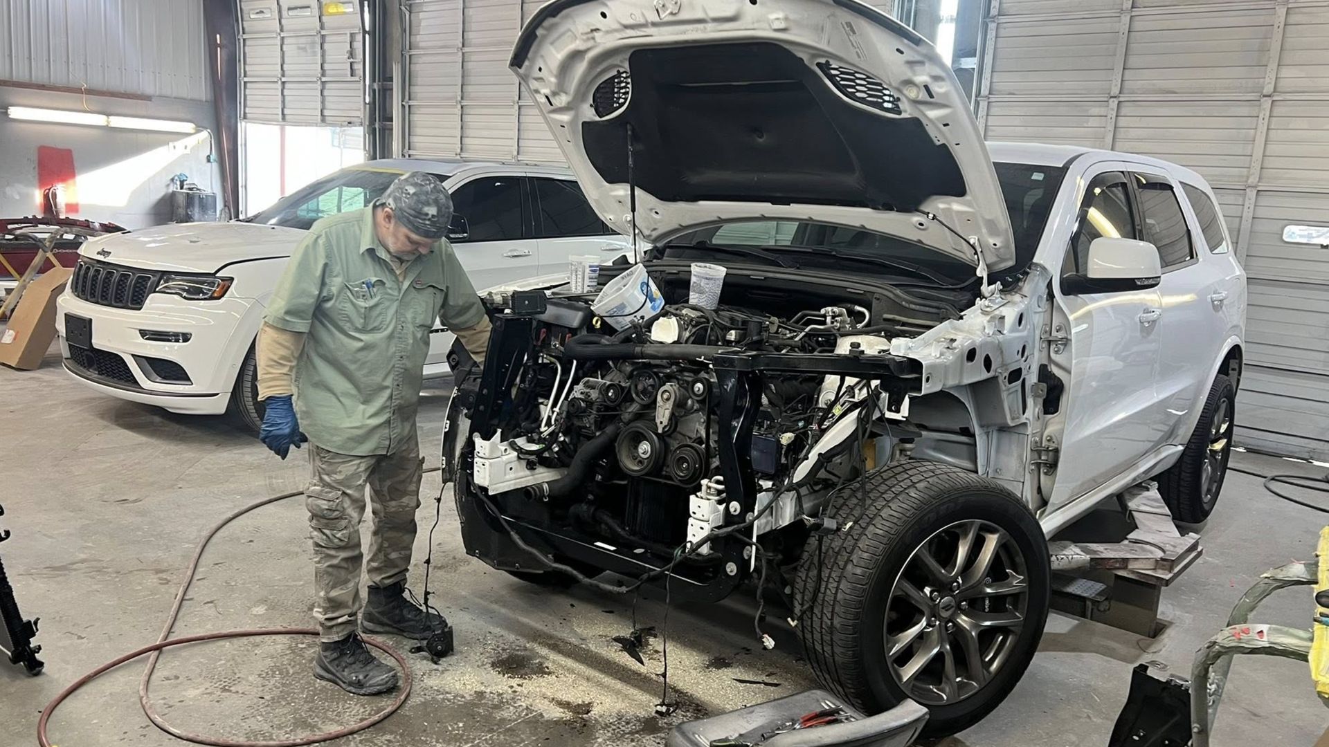 A mechanic works on the front engine area of a white SUV with its hood open in a repair shop.