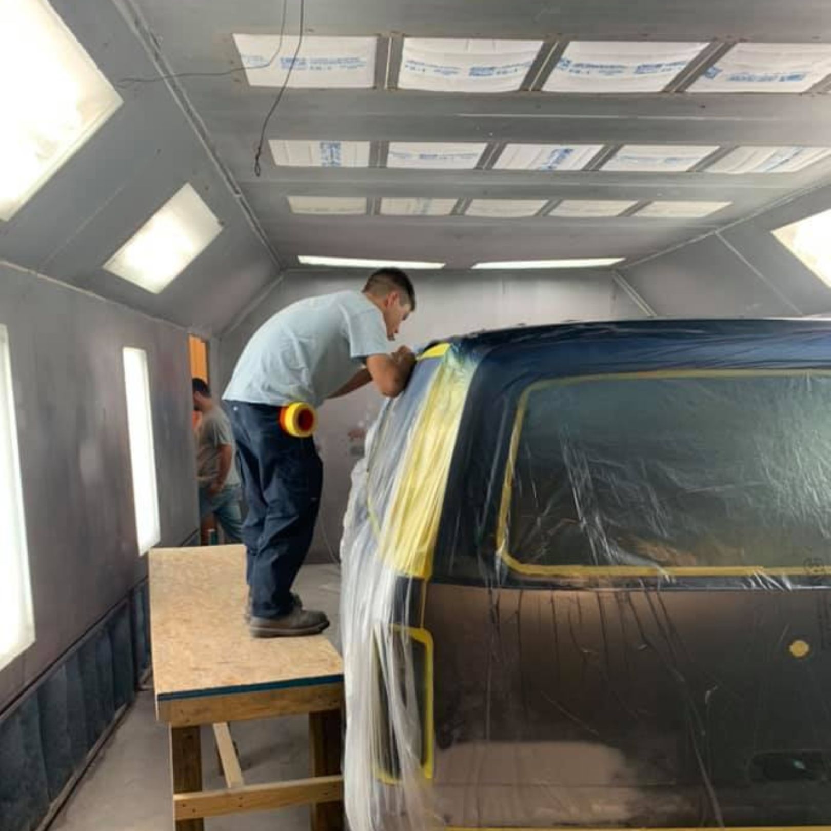 A worker stands on a wooden platform inside a spray booth, preparing a vehicle covered in plastic sheeting for painting.