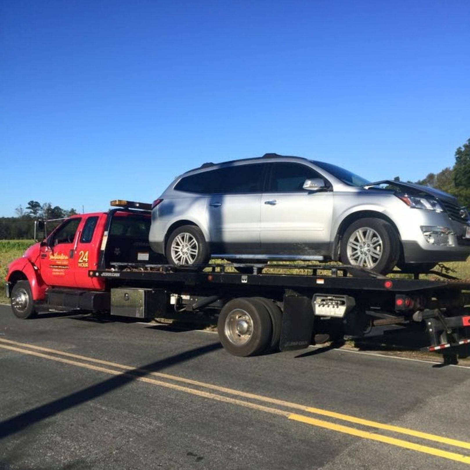 A red flatbed tow truck transporting a silver SUV on a paved road under a clear blue sky.