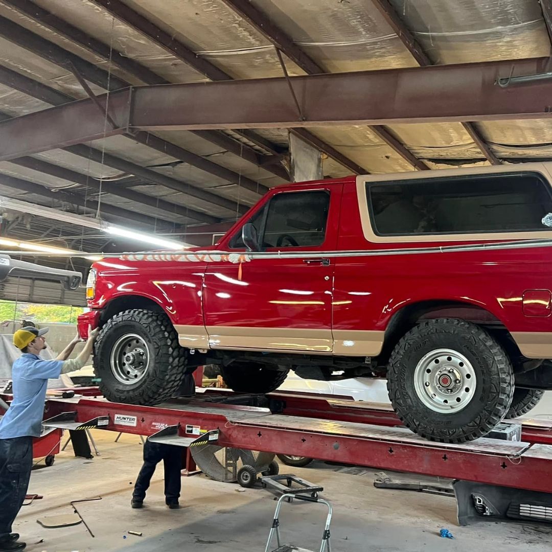 A red and tan vintage SUV is elevated on a lift in an auto shop while a technician works on the undercarriage.