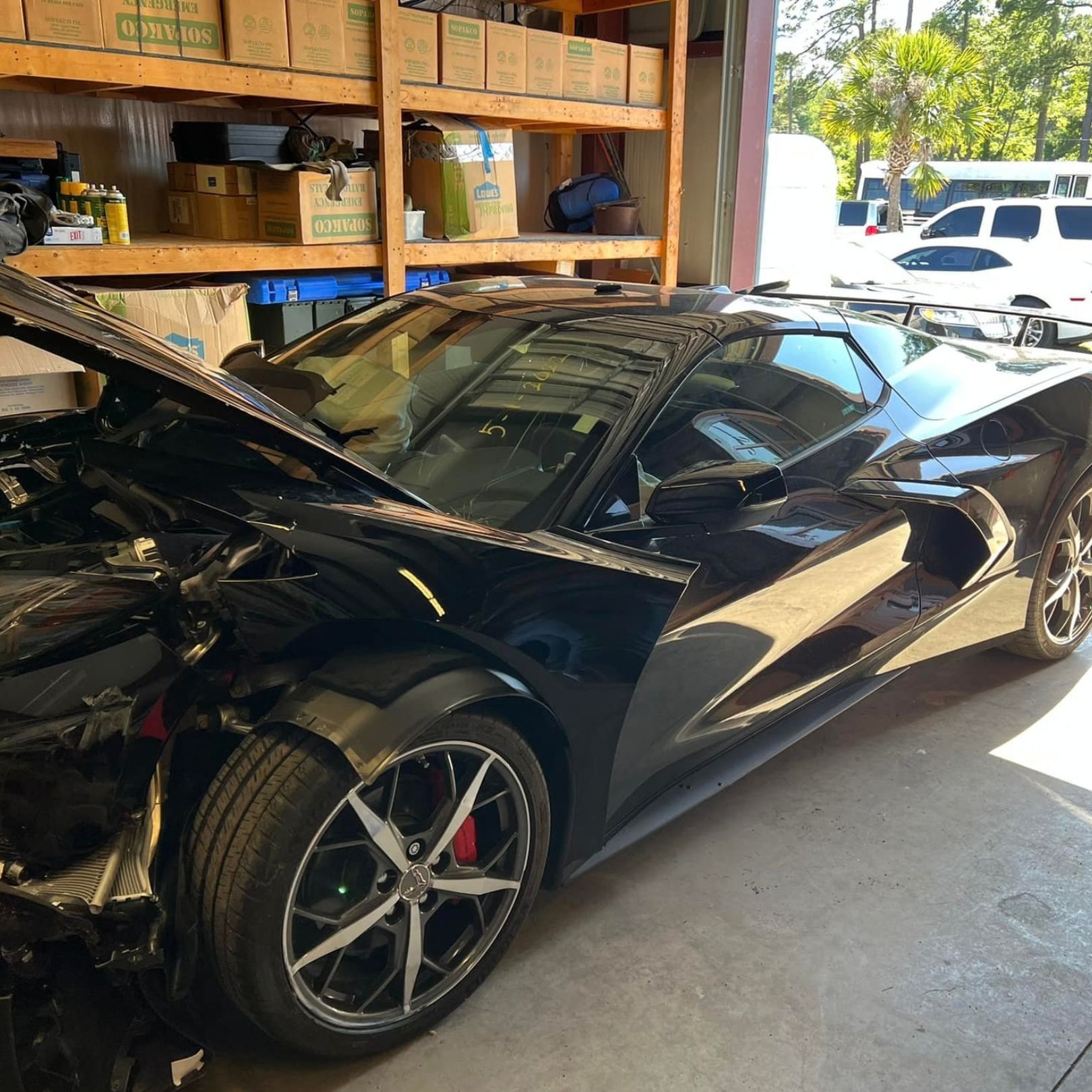 A black Chevrolet Corvette C8 with a damaged front end sits inside a warehouse filled with shelving units.