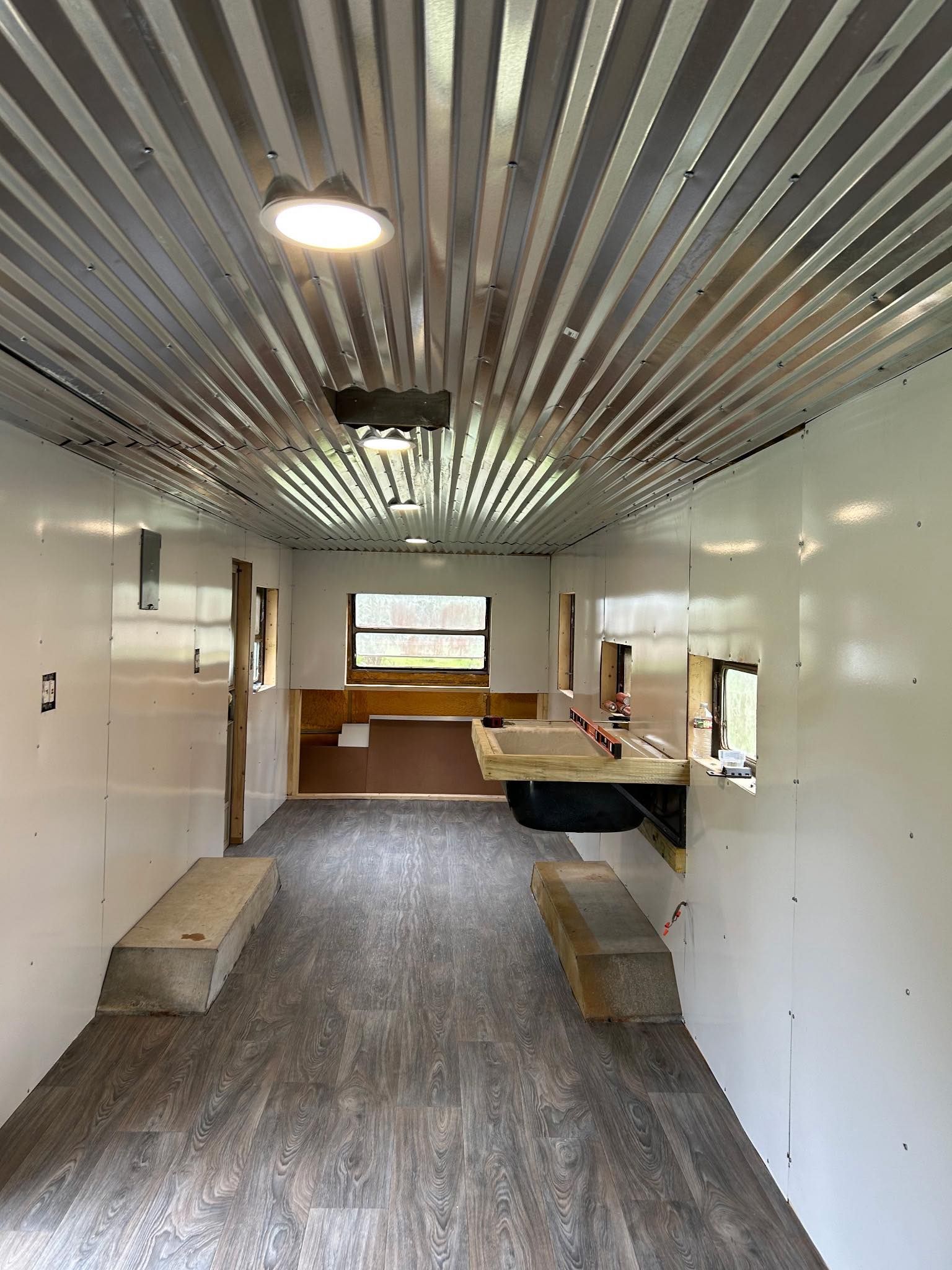 Interior of a trailer with white walls, wood-look flooring, a corrugated metal ceiling, and built-in wooden benches.