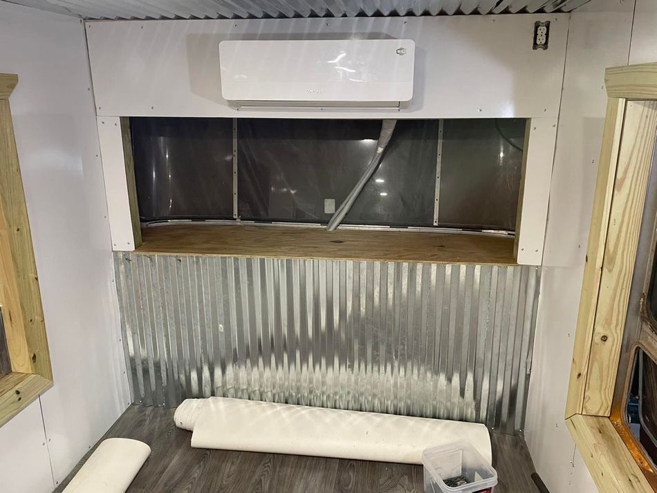 Interior view of a room under construction with corrugated metal siding, a wood counter, and a wall-mounted AC unit.