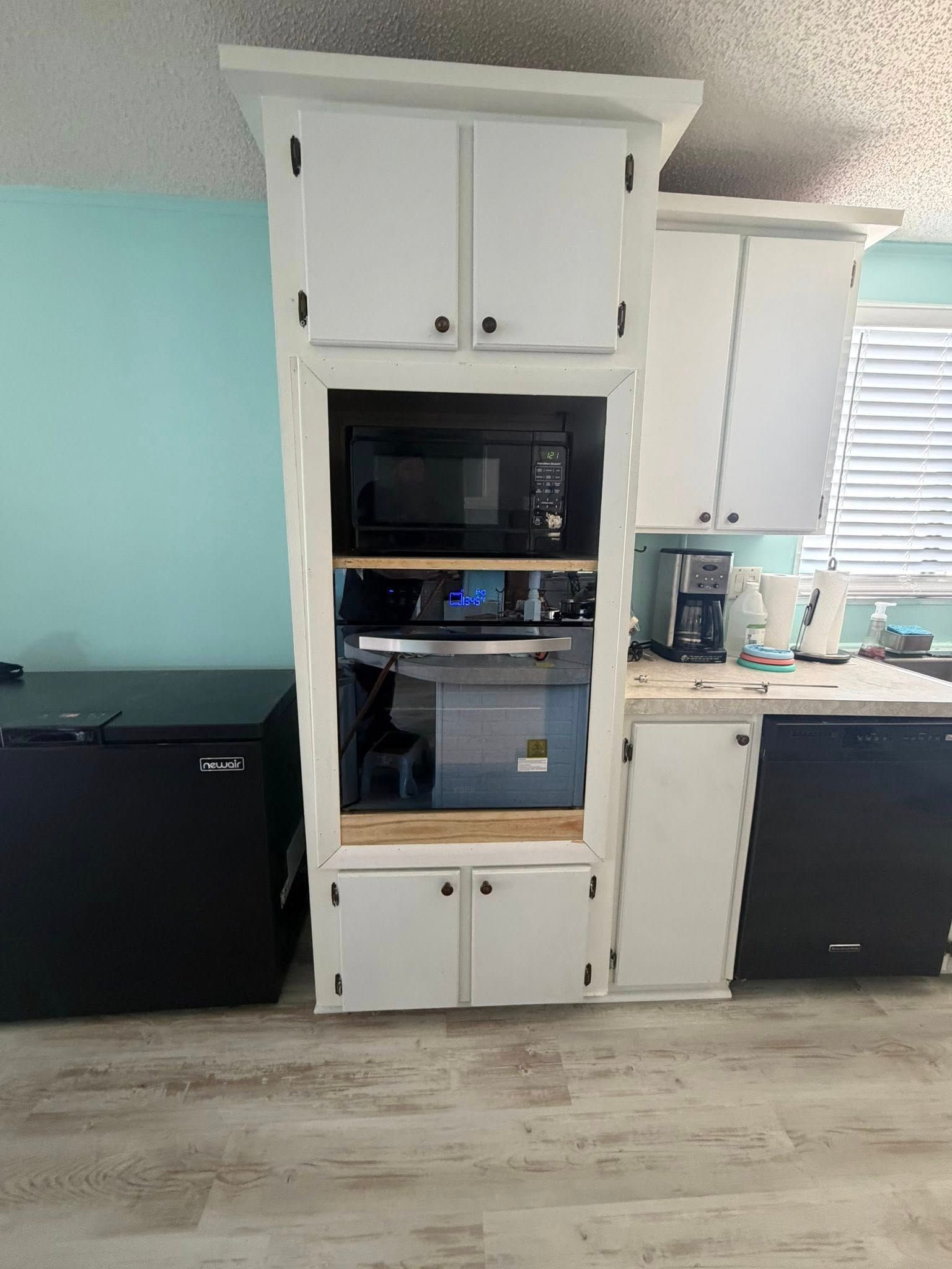 White kitchen cabinet with built-in oven and drawers between a black fridge and a sink under a window.