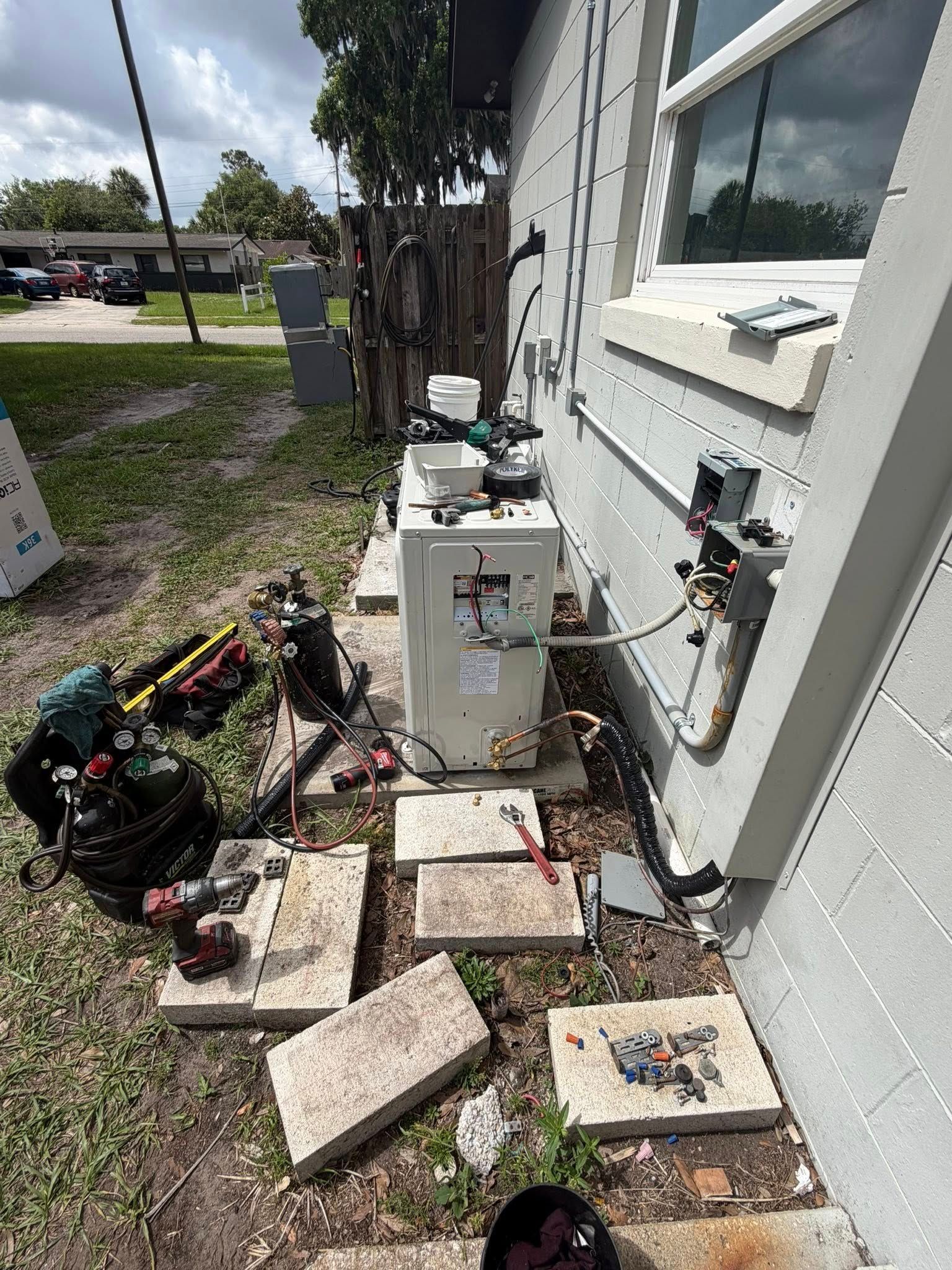 Outdoor utility area beside a house with scattered pavers, tools, and electrical equipment on the ground.