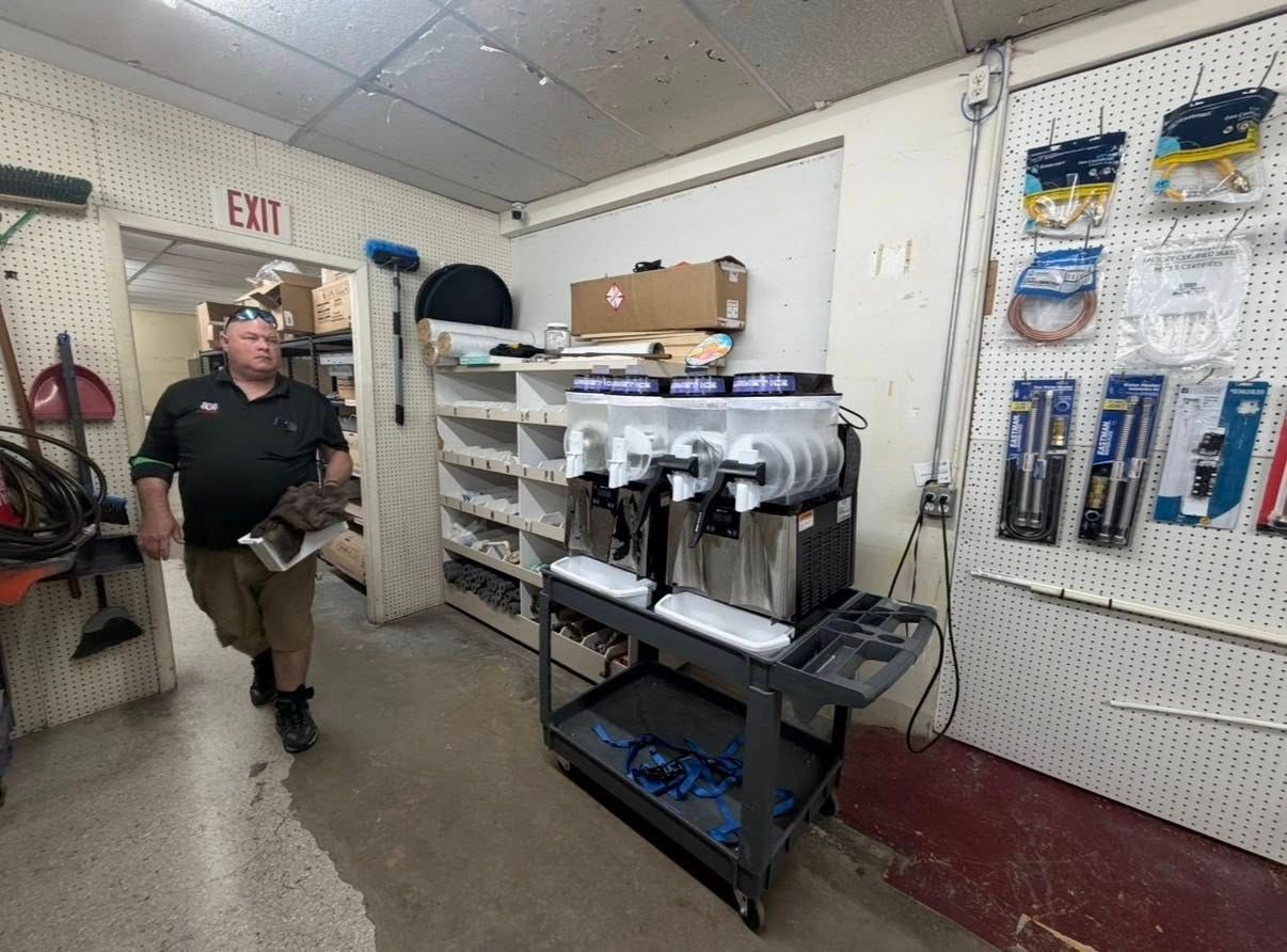 Man in a workshop beside a pegboard wall, rolling cart, and stacked supplies under an EXIT sign