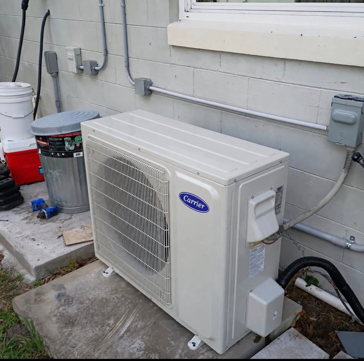 Outdoor HVAC air conditioner unit beside a house wall with pipes and a white bucket nearby