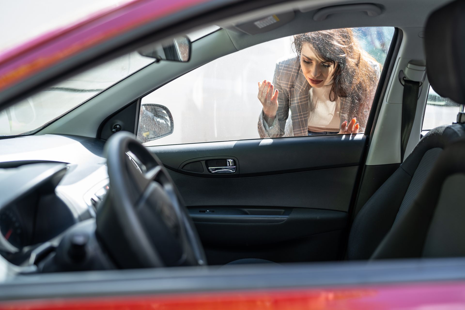 A woman is looking through the window of a locked car.