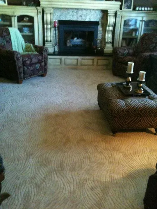 Living room with a fireplace, two armchairs, a patterned ottoman, and light-colored patterned carpet.