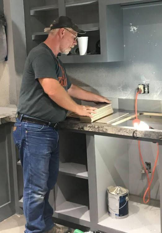 Man in a baseball cap placing tiles on a countertop in a kitchen renovation. Gray cabinets and walls, light-colored tiles.