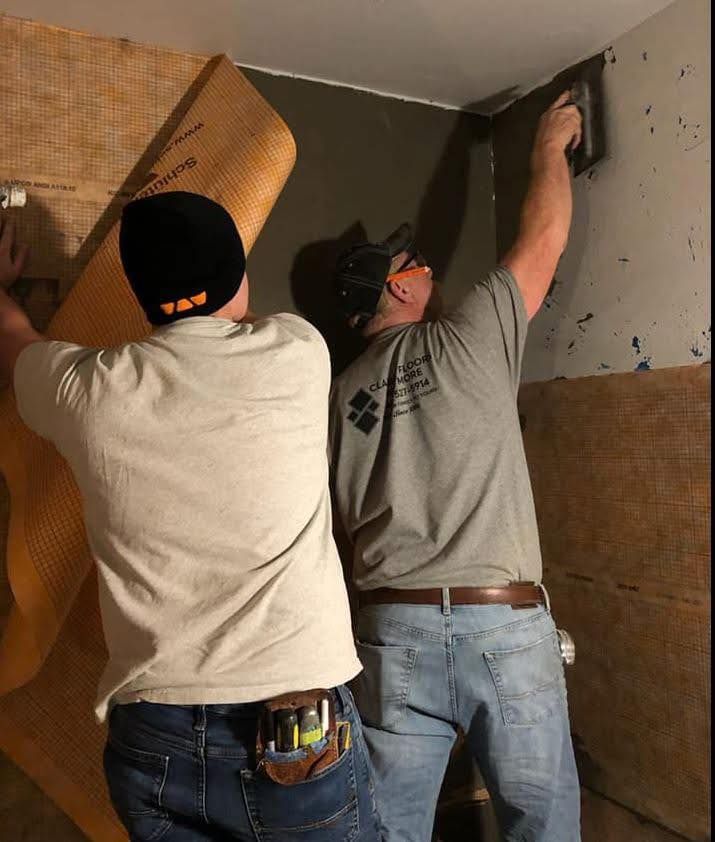 Two construction workers installing wall tile in a room. One worker is applying mortar, the other is holding up a membrane.