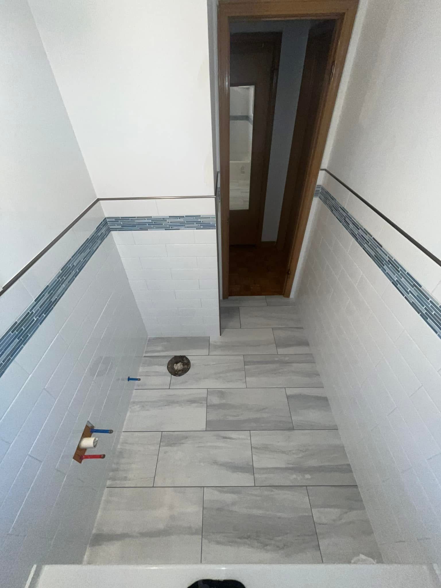 A bathroom with newly tiled walls and floor in shades of white and gray. A wooden door is visible at the end of the space.