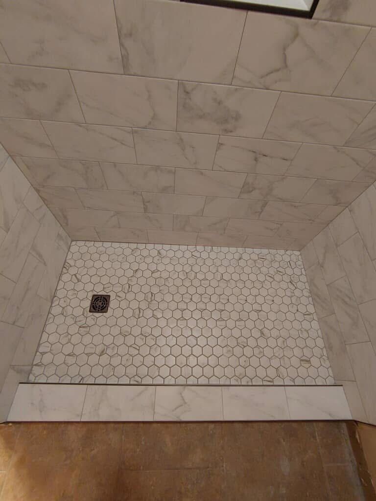 A shower stall with white marble tile walls and a hexagon tile floor, a drain, and a curb at the bottom.