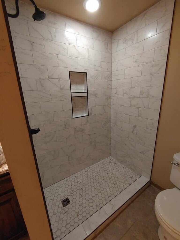 A newly tiled shower with white marble-look walls, a hexagon-tiled floor, and a built-in shelf.