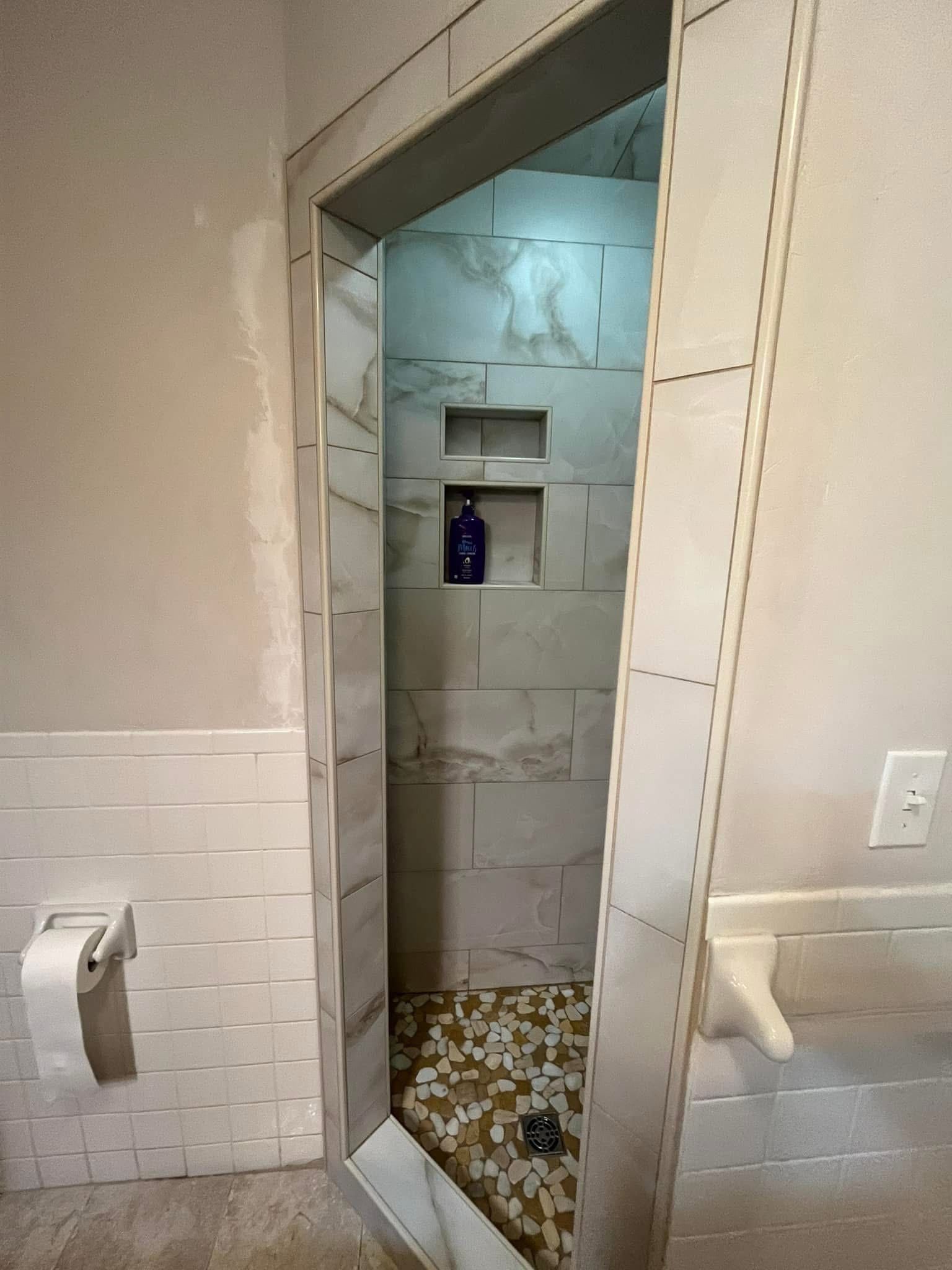 Narrow shower with tiled walls and floor, accessible via an opening. A toilet paper holder is visible on the left.