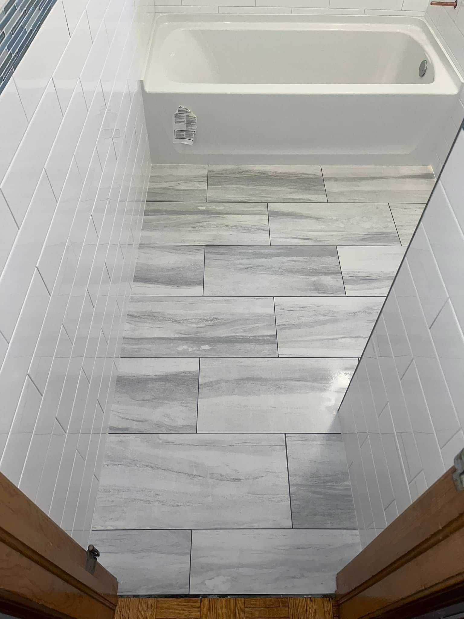 Bathroom with a white bathtub and gray and white patterned floor tiles. White tile walls with small, rectangular patterns.
