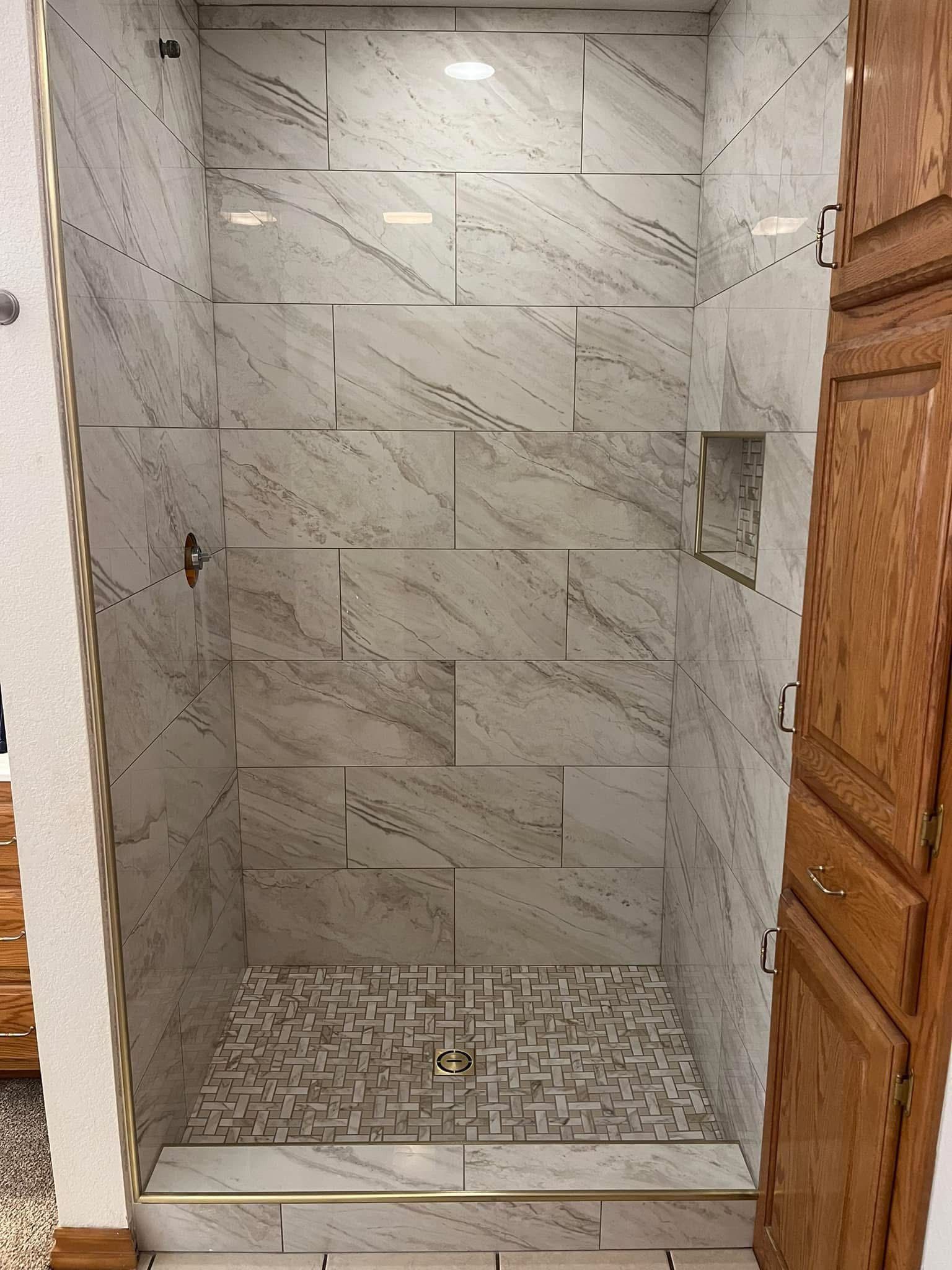 A tiled shower with a mosaic floor and built-in shelving. Light gray marble-like tiles cover the walls, and there 