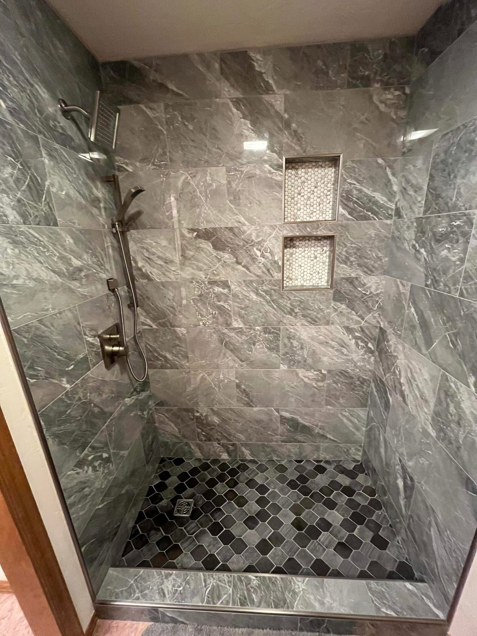 A modern shower with gray marbled tiles and a mosaic tile floor. Two recessed shelves and a showerhead are visible.