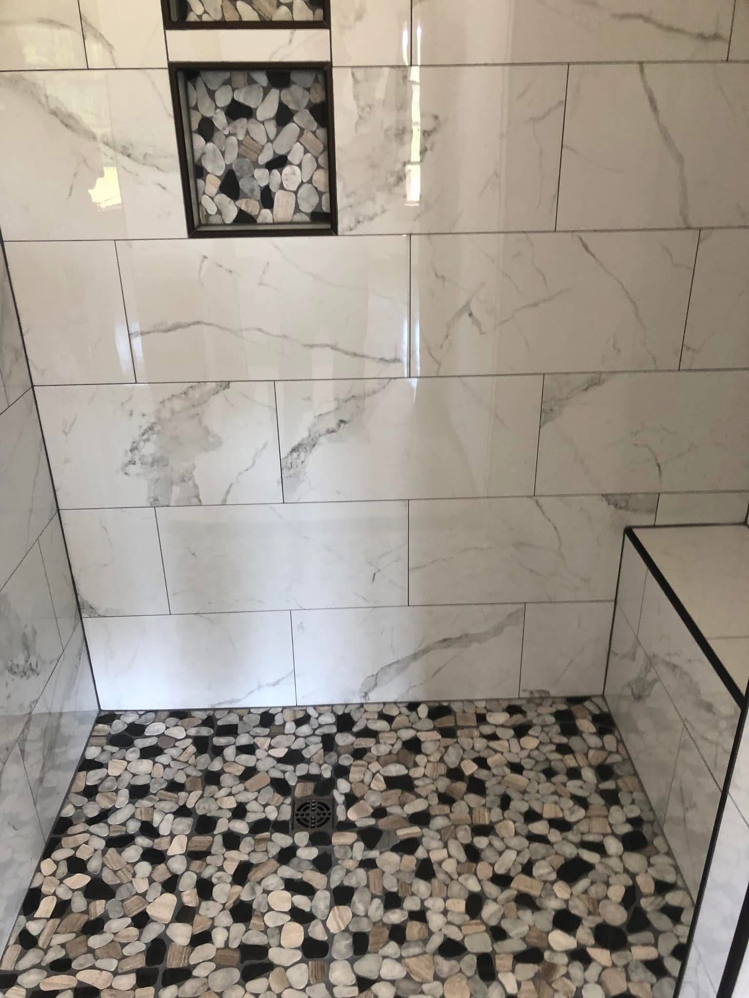 White and gray tiled shower with a pebble stone floor. A recessed shelf holds decorative stones.