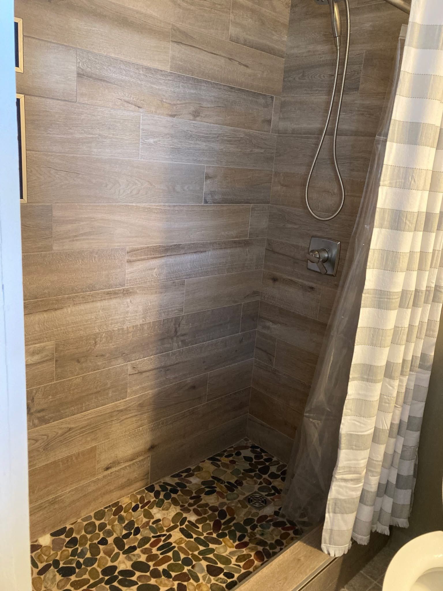 A shower with tan-colored, wood-like tile walls and a pebble floor. A white and gray striped shower curtain hangs
