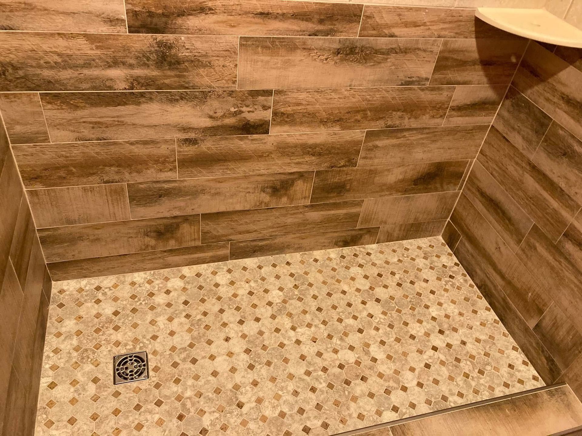 Shower with brown wood-look tile walls and small beige mosaic floor tiles. A small corner shelf is visible.