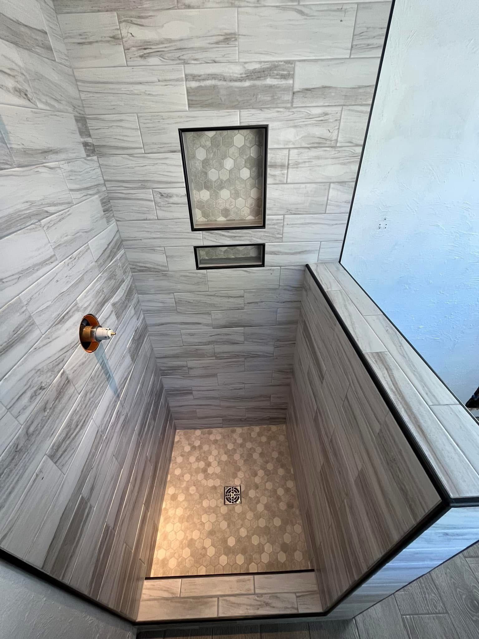 A shower interior with gray and white tiled walls and floor. A recessed shelf and faucet are visible.