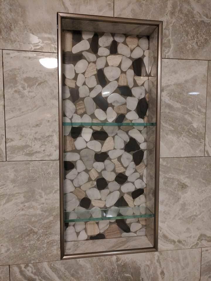 A recessed shower niche with a pebble tile backing and glass shelves, framed in silver. The surrounding tile is gray 