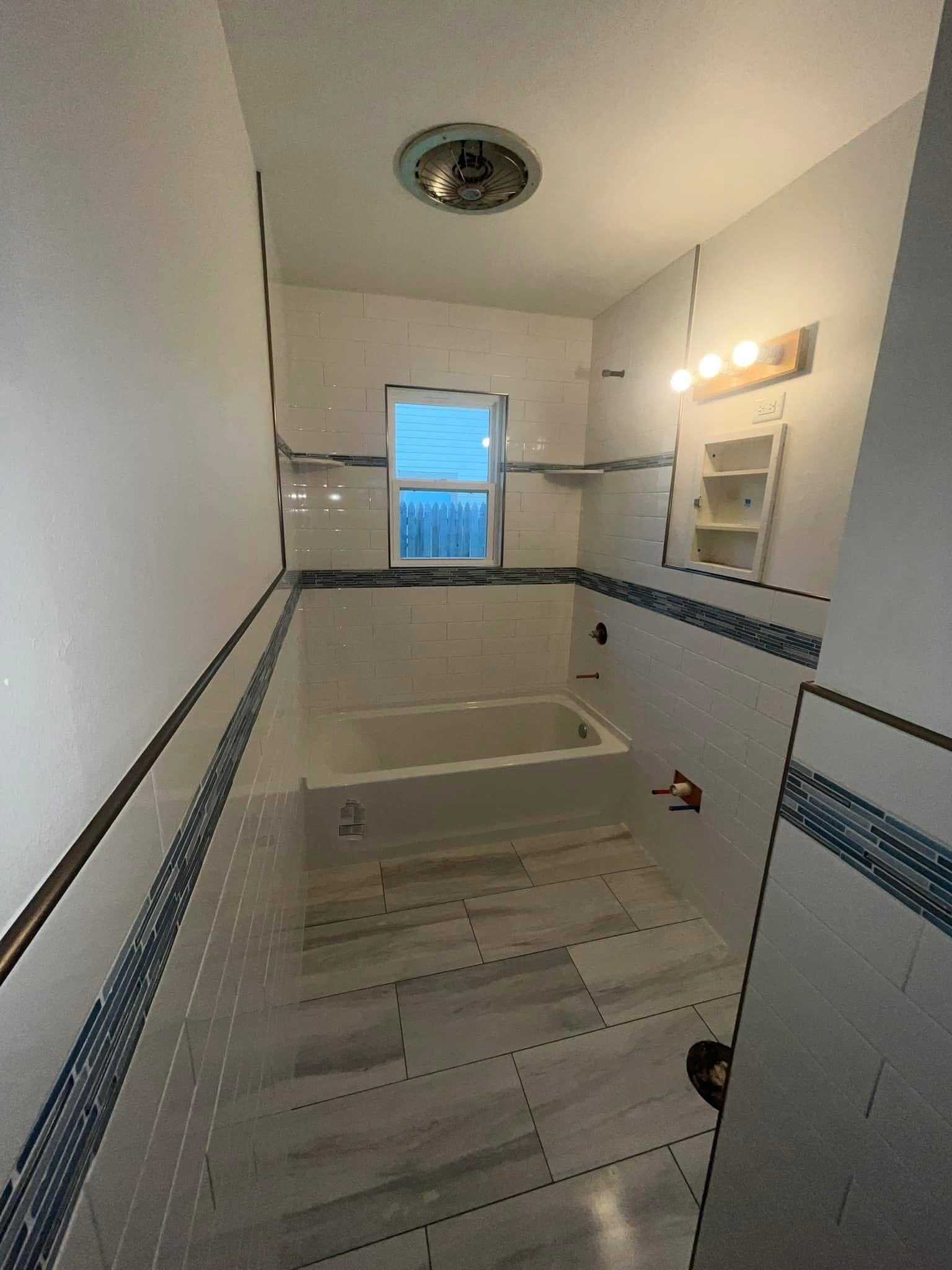 Narrow bathroom with white and blue tiled walls, a bathtub, and a window with a view.