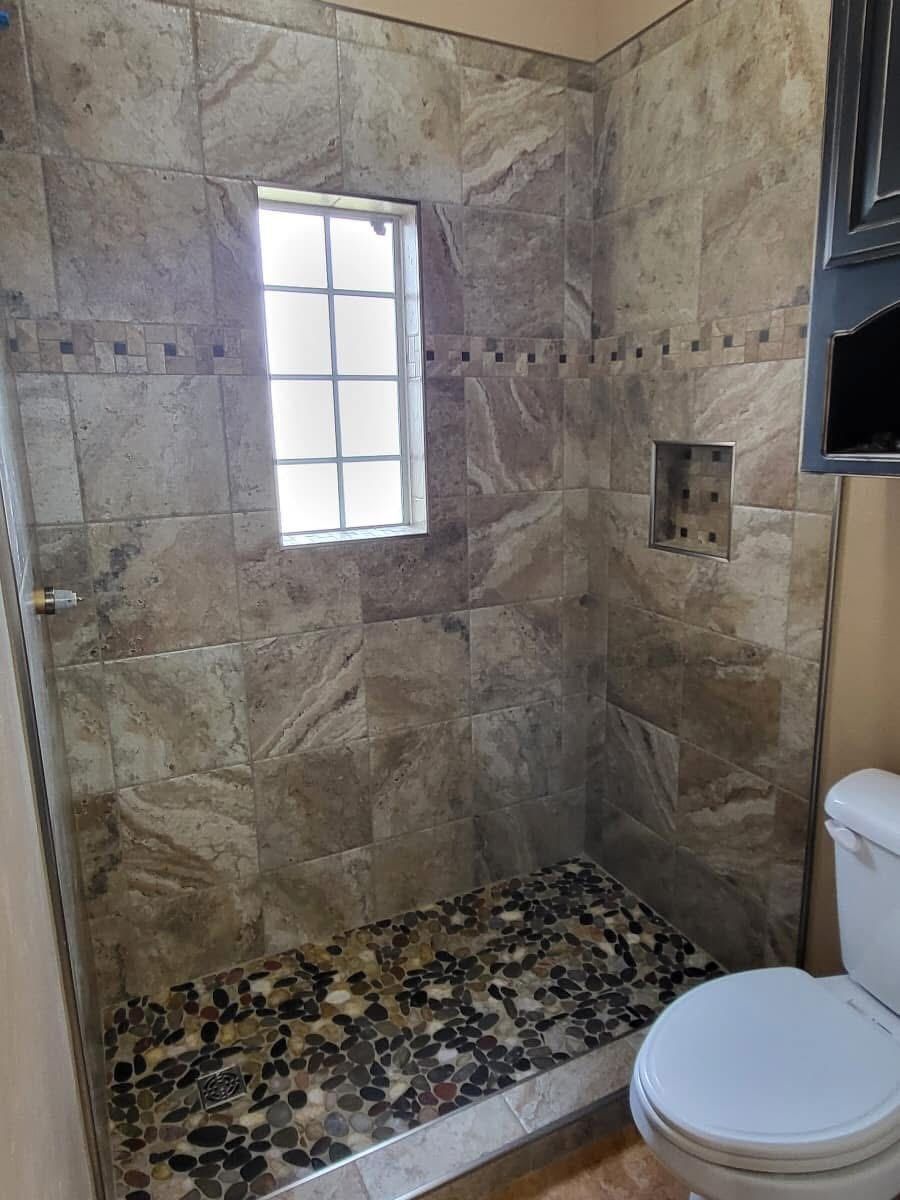 Shower stall with patterned tile walls and a pebble floor, with a window and recessed shelf. A white toilet sits adjacent.
