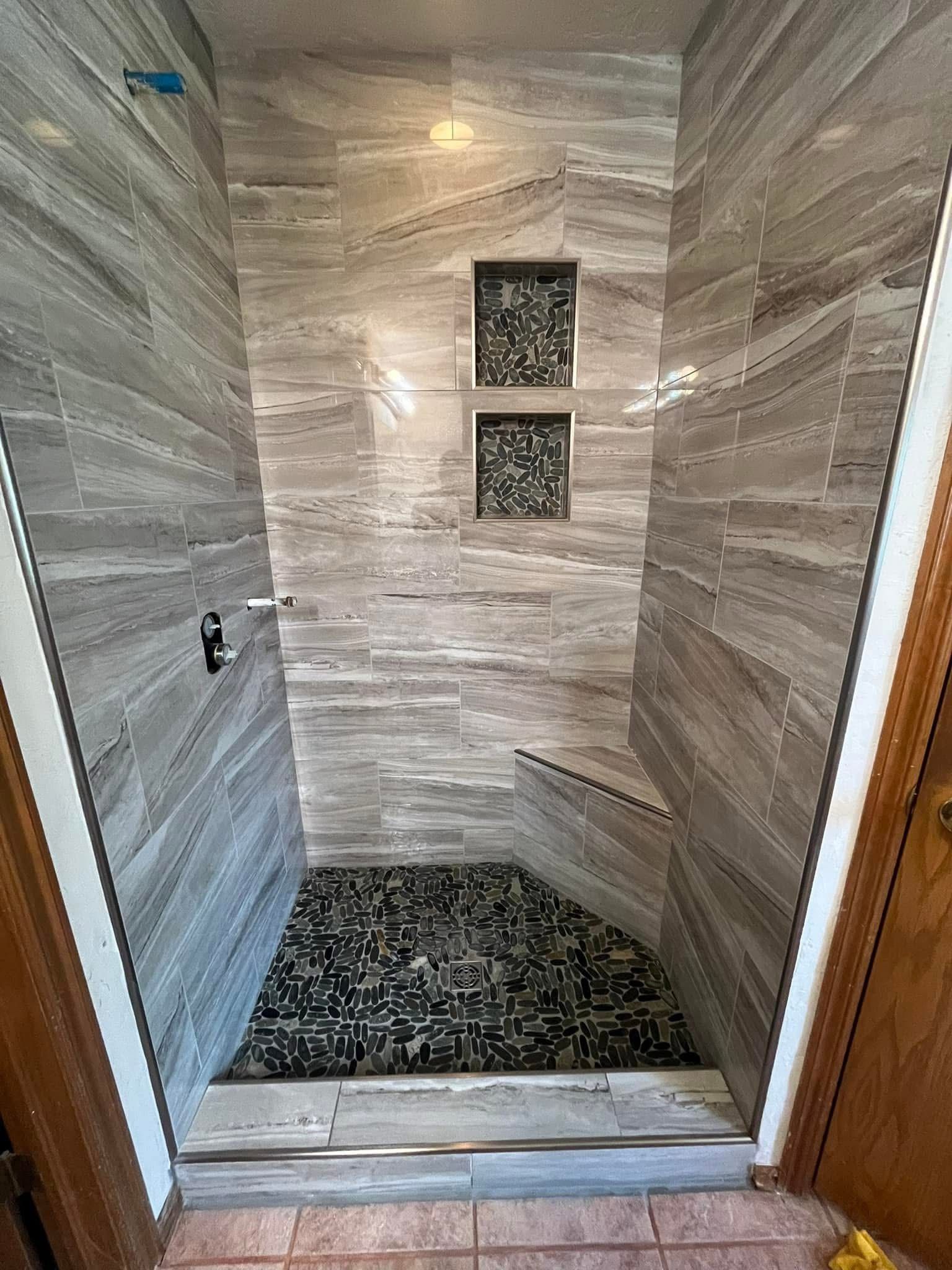 Newly tiled walk-in shower with gray stone-look walls and black and gray pebble floor. Two inset shelves, silver fixtures.