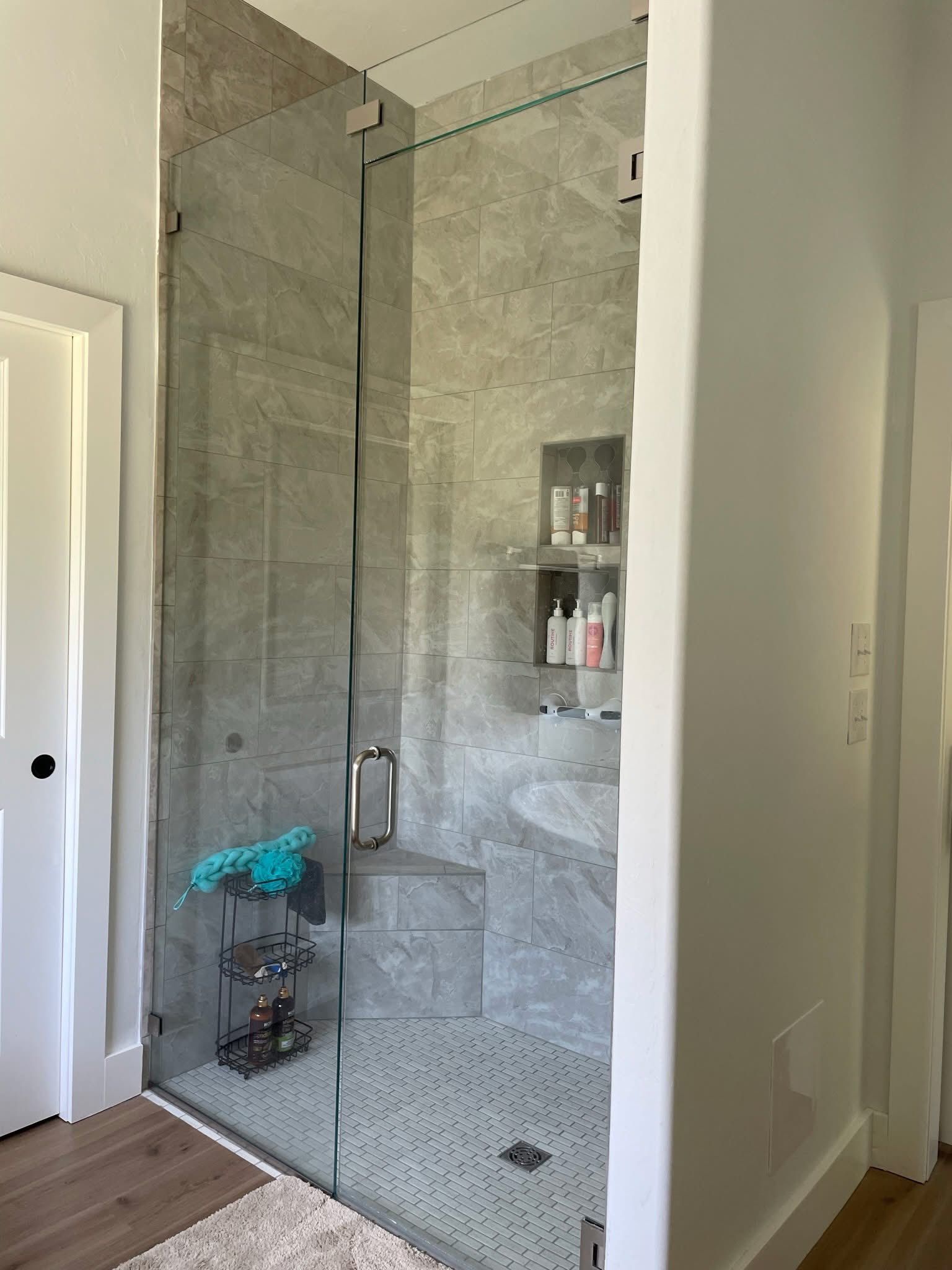 A glass shower enclosure with gray tile walls and pebble floor. Inside, there are shelves with toiletries and a shower head.