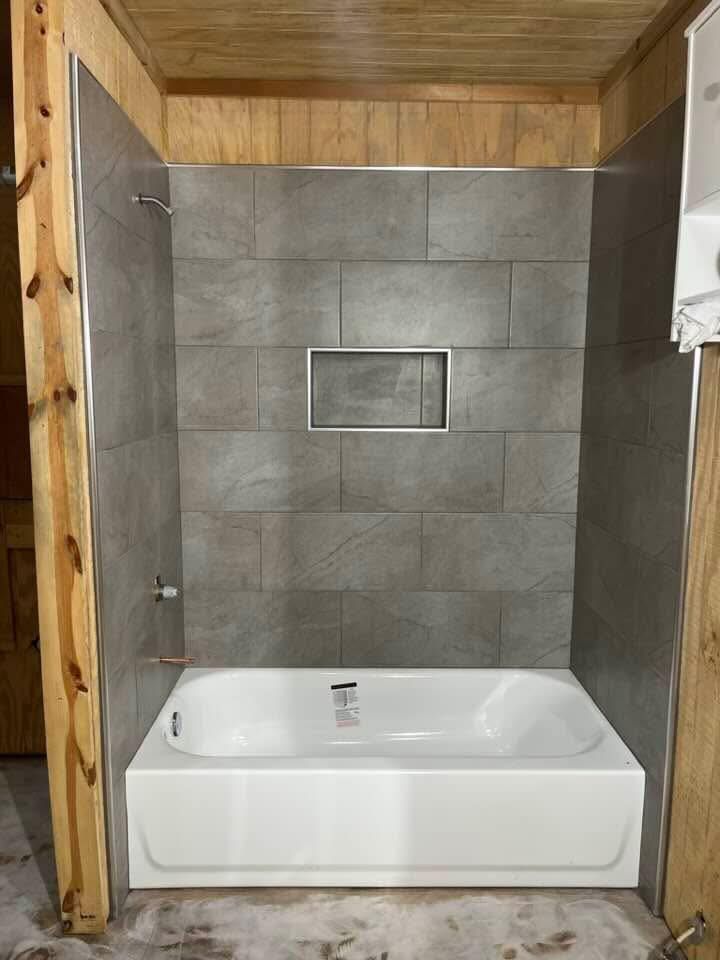Bathroom with a white tub, grey tiled shower walls, and a recessed shelf. The shower is framed by wooden beams.