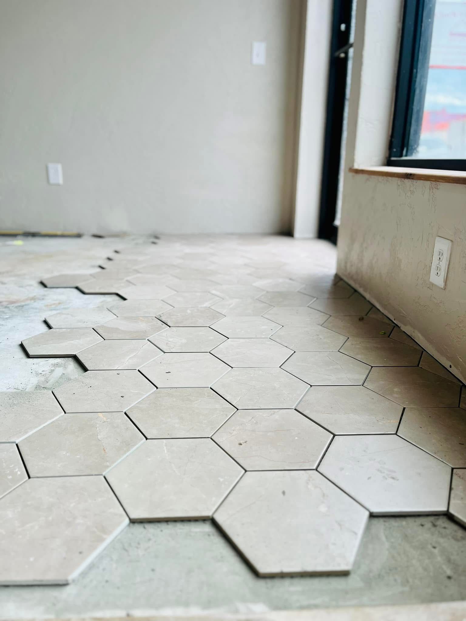Hexagon tiles being laid on a concrete floor in a room under construction. Beige tiles are arranged in a honeycomb pattern.