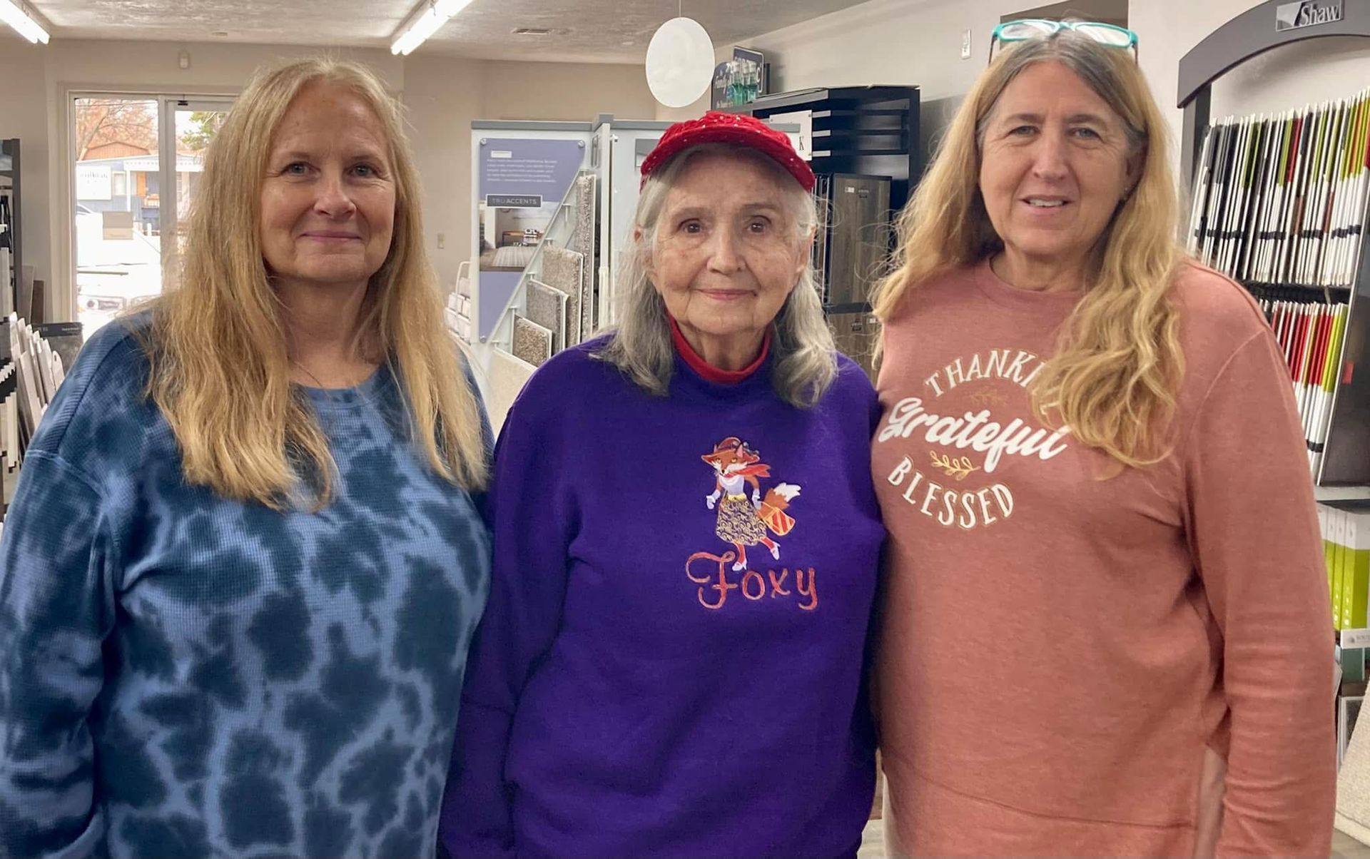 Three women smiling, standing indoors in a store. Woman in the center wears a red hat and purple sweatshirt, 