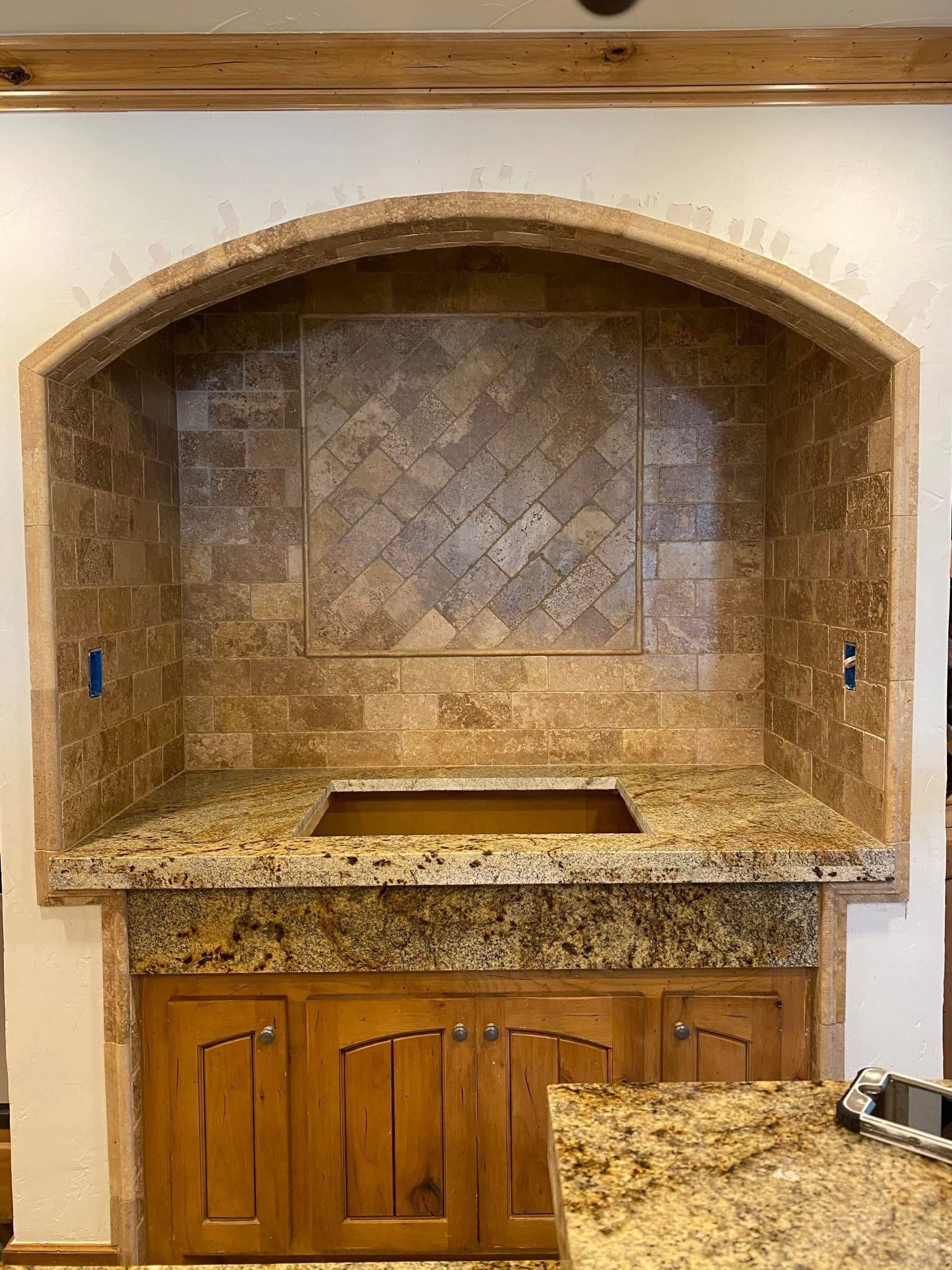 A kitchen alcove with a granite countertop, tiled backsplash, and wooden cabinets. The archway above is also tiled.