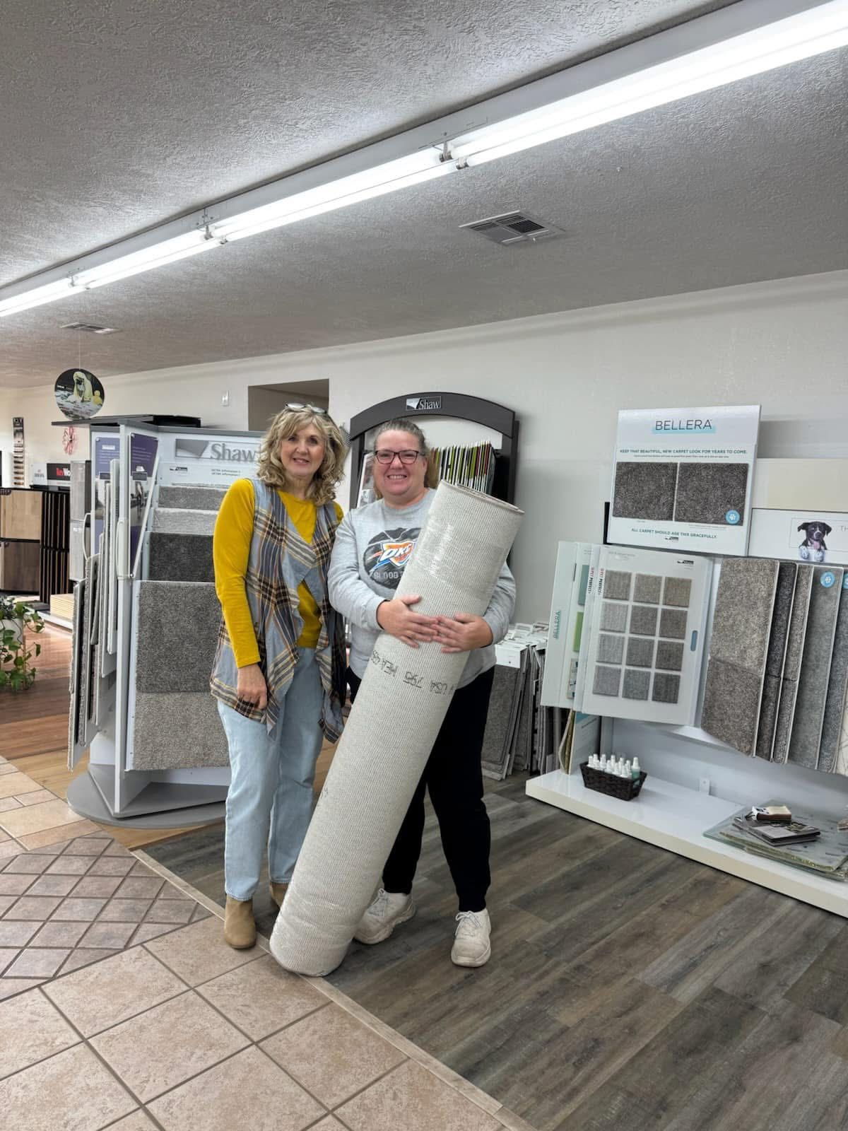 Two women in a flooring store; one holds a rolled-up carpet, smiling. Various flooring samples displayed on stands.