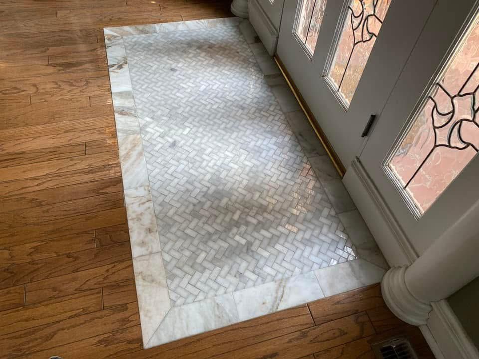 Herringbone tile inset at a doorway, bordered by a light-colored stone. The tile is on a wood floor next to a white door.