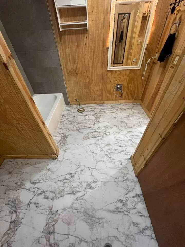 A small bathroom with marble-patterned flooring and wood-paneled walls. A bathtub, mirror, and fixtures are visible.