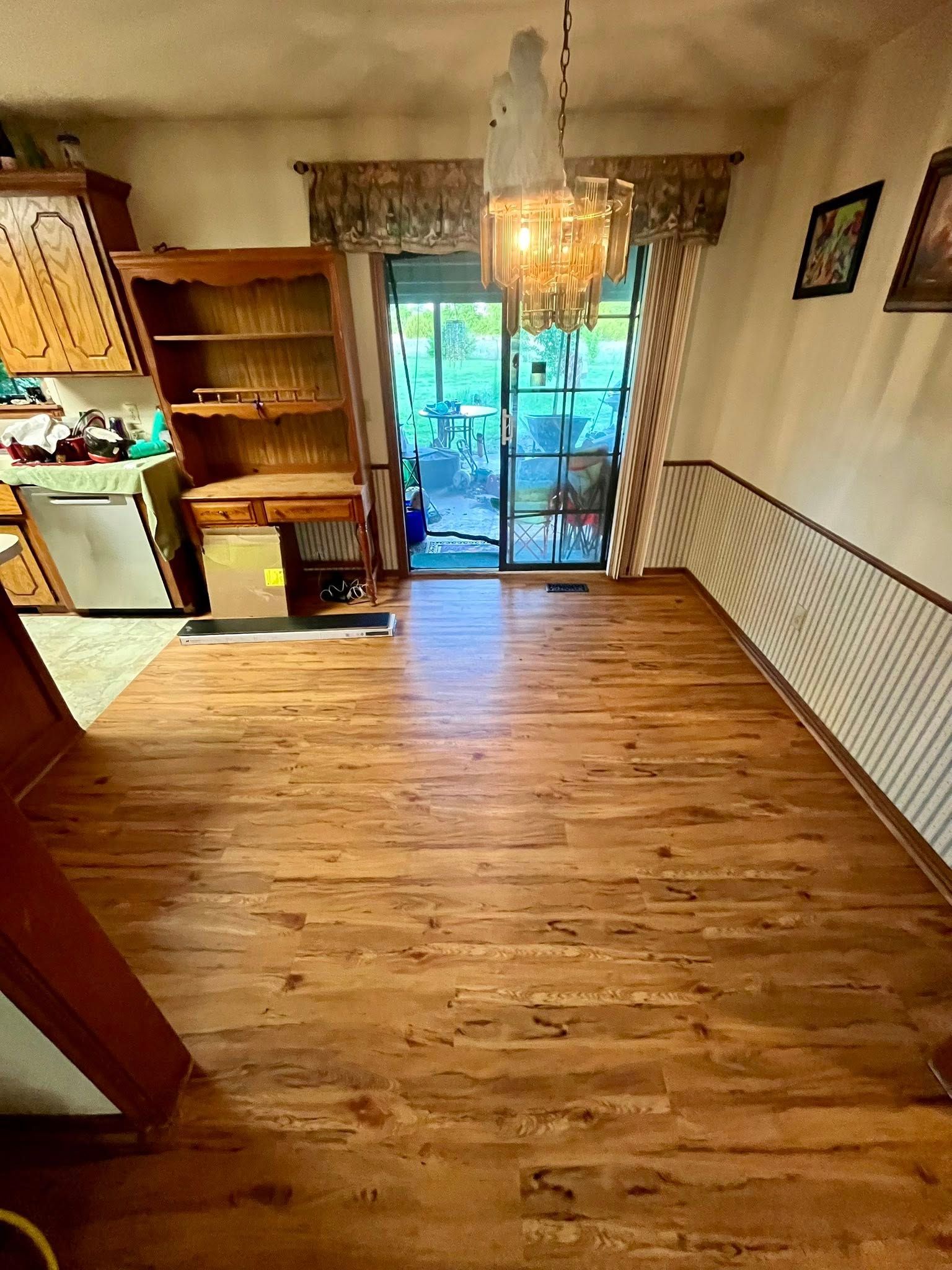 Wooden floor extends from a kitchen towards a sliding glass door. chandelier hangs above, with cabinets