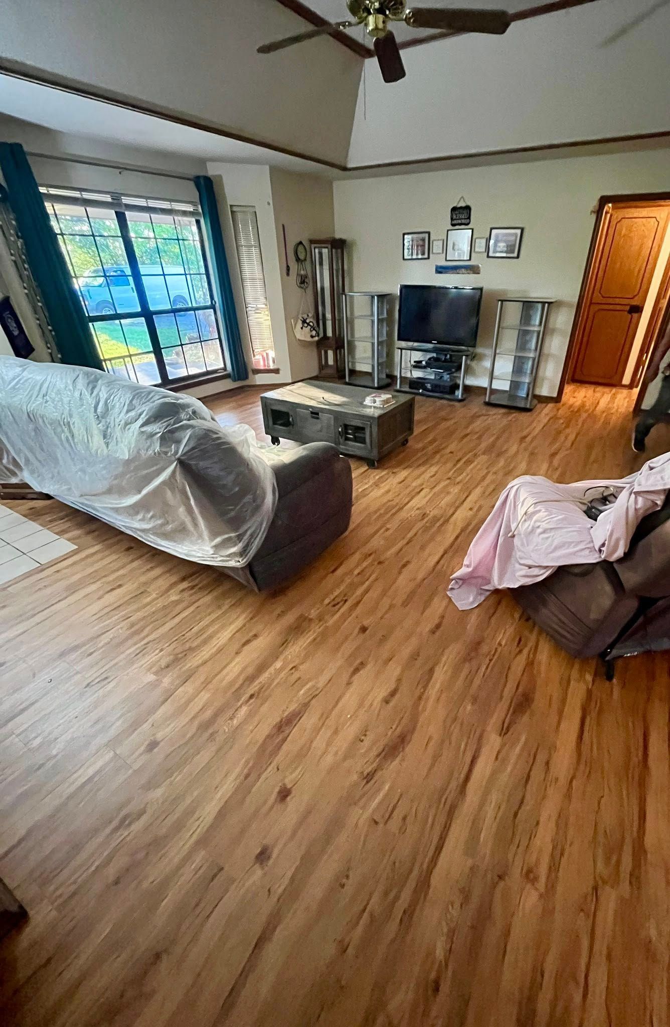 Living room with wood flooring, couches, a TV, and shelves. Sunlight streams in from the large window 