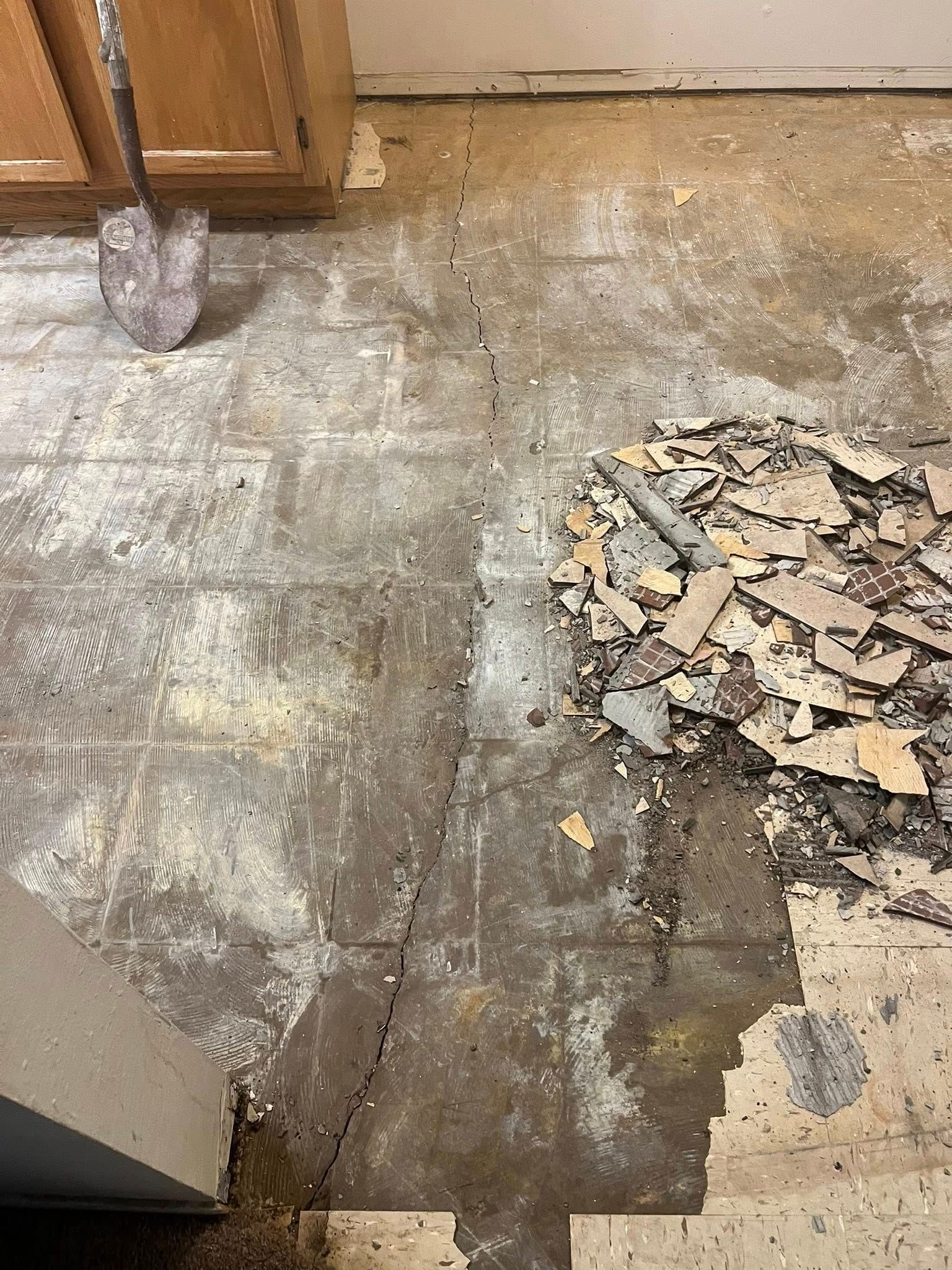 A kitchen floor with ripped-up flooring and debris. A shovel leans against a cabinet.