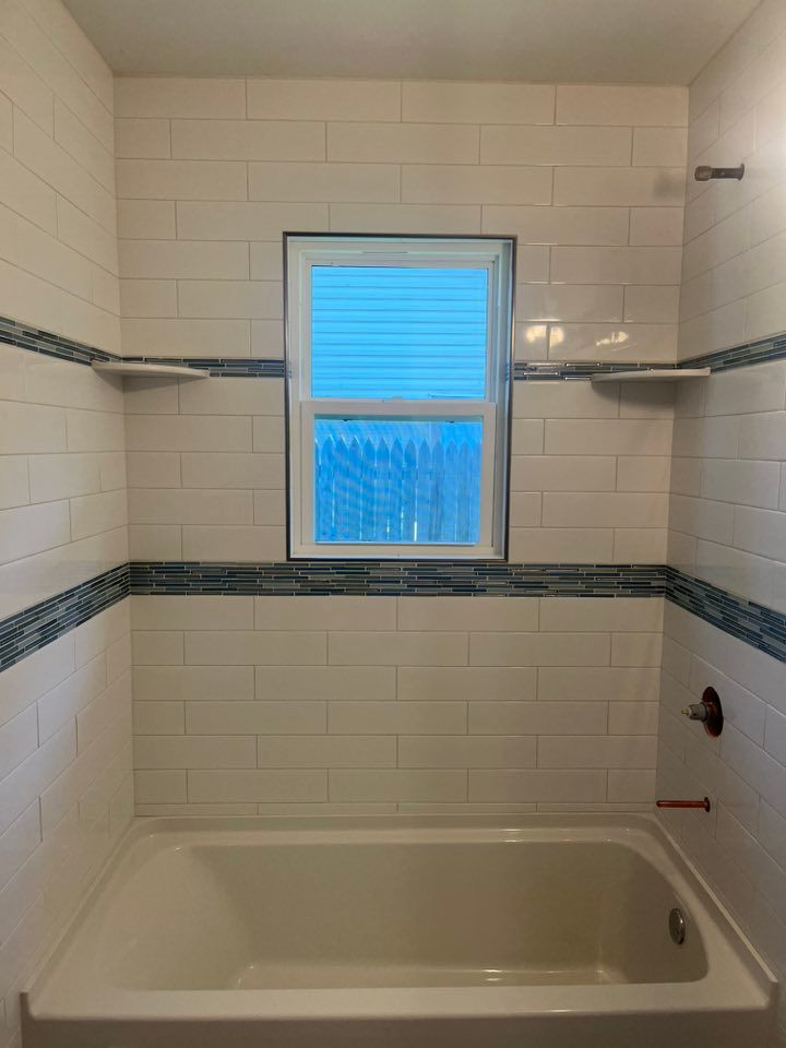 Bathroom with white and blue tiled walls, a window, and a bathtub. Two shelves flank the window, and a showerhead is visible.
