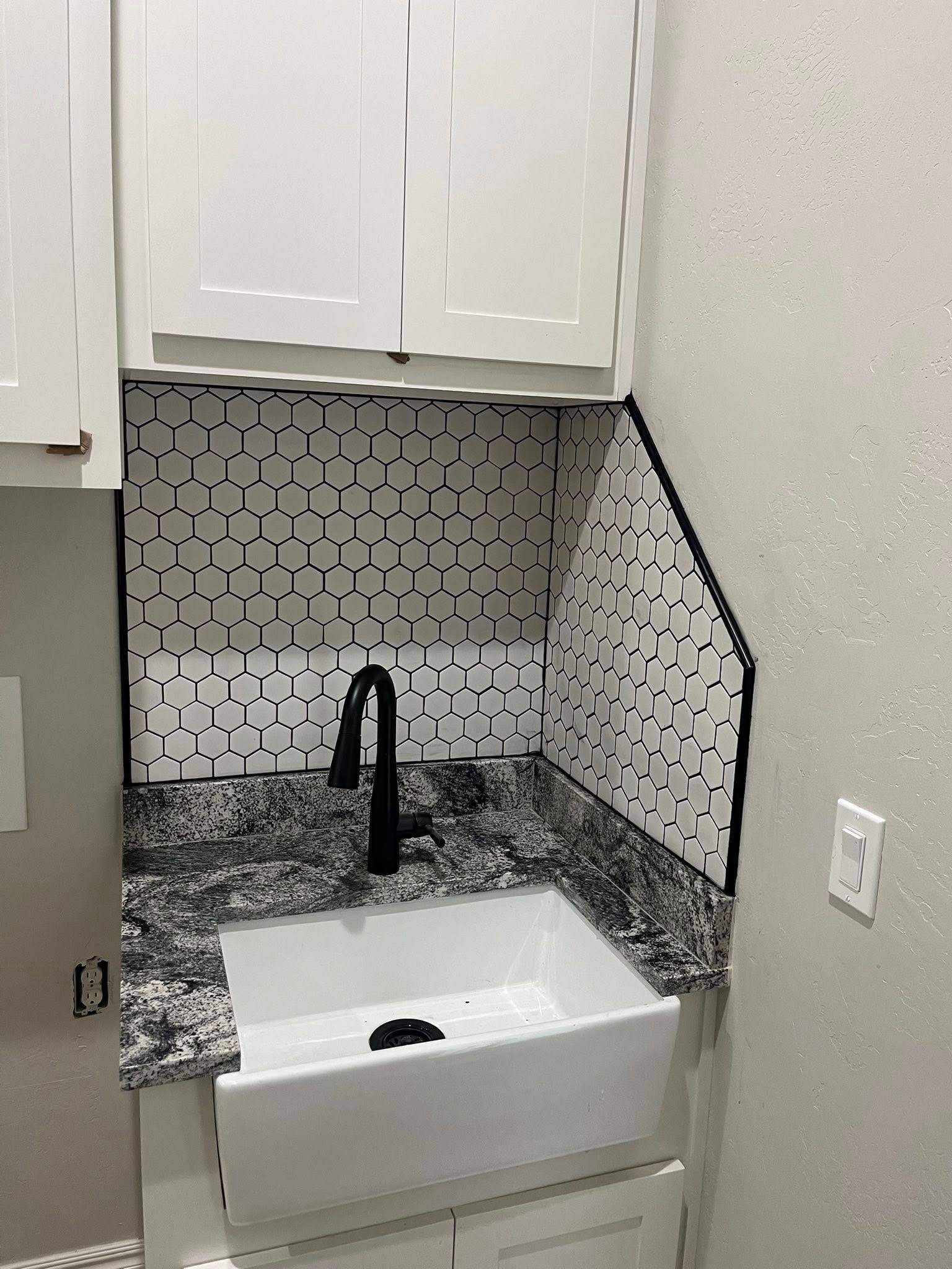 A utility sink area with white cabinets, granite countertop, black faucet, and patterned tile backsplash.
